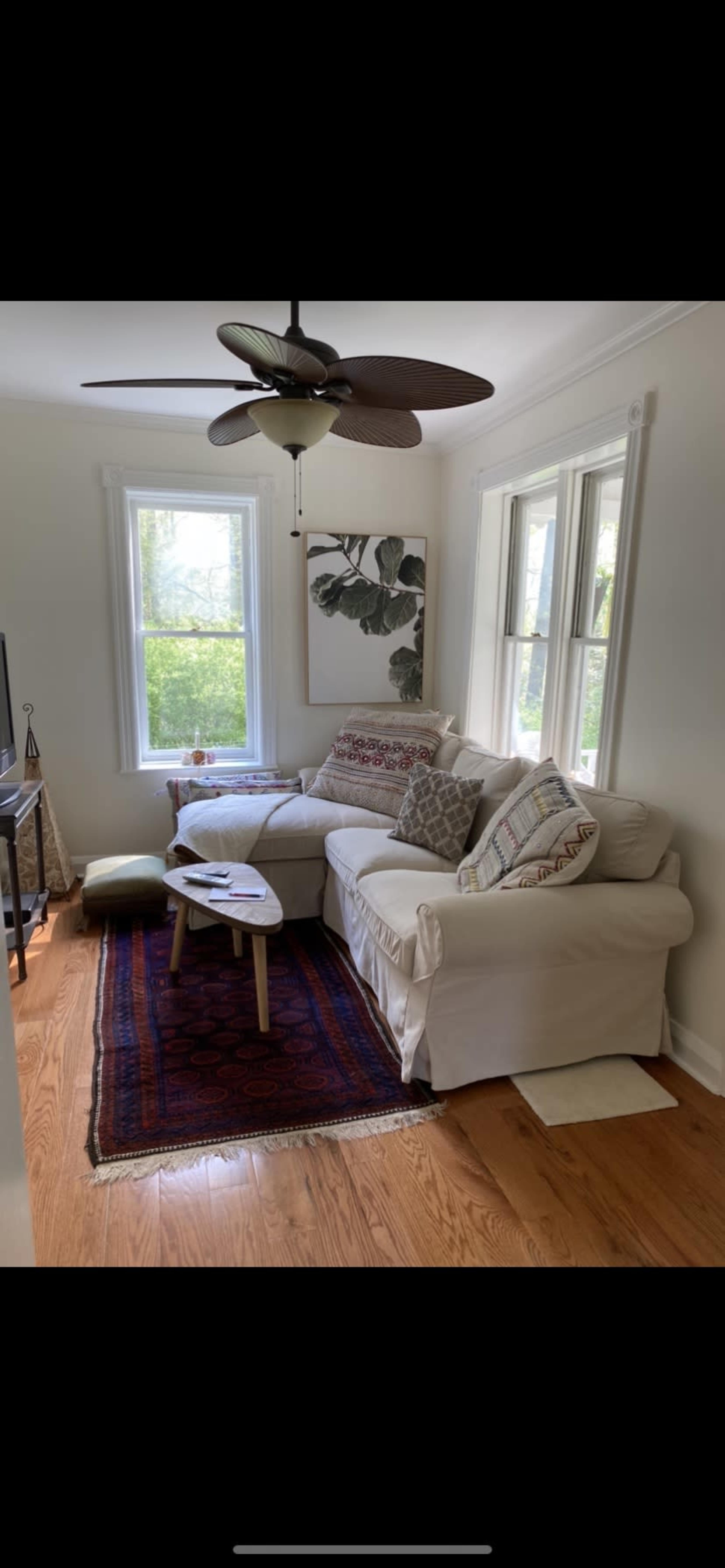 The image shows a living room with a white sectional sofa, a small wooden table, and large windows allowing natural light to enter.