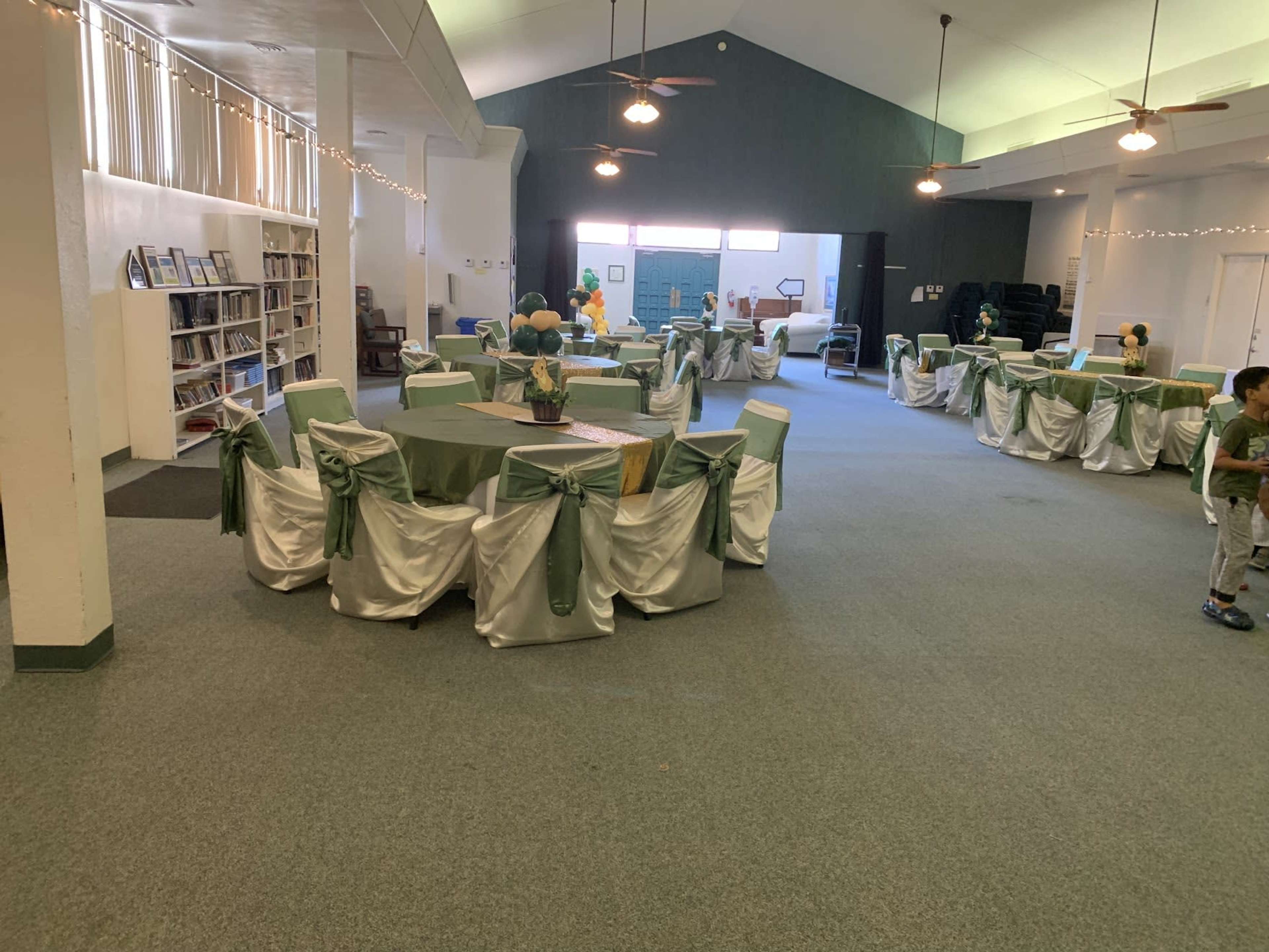 The room features multiple tables set with green tablecloths and white chair covers, with a library shelf visible in the background.
