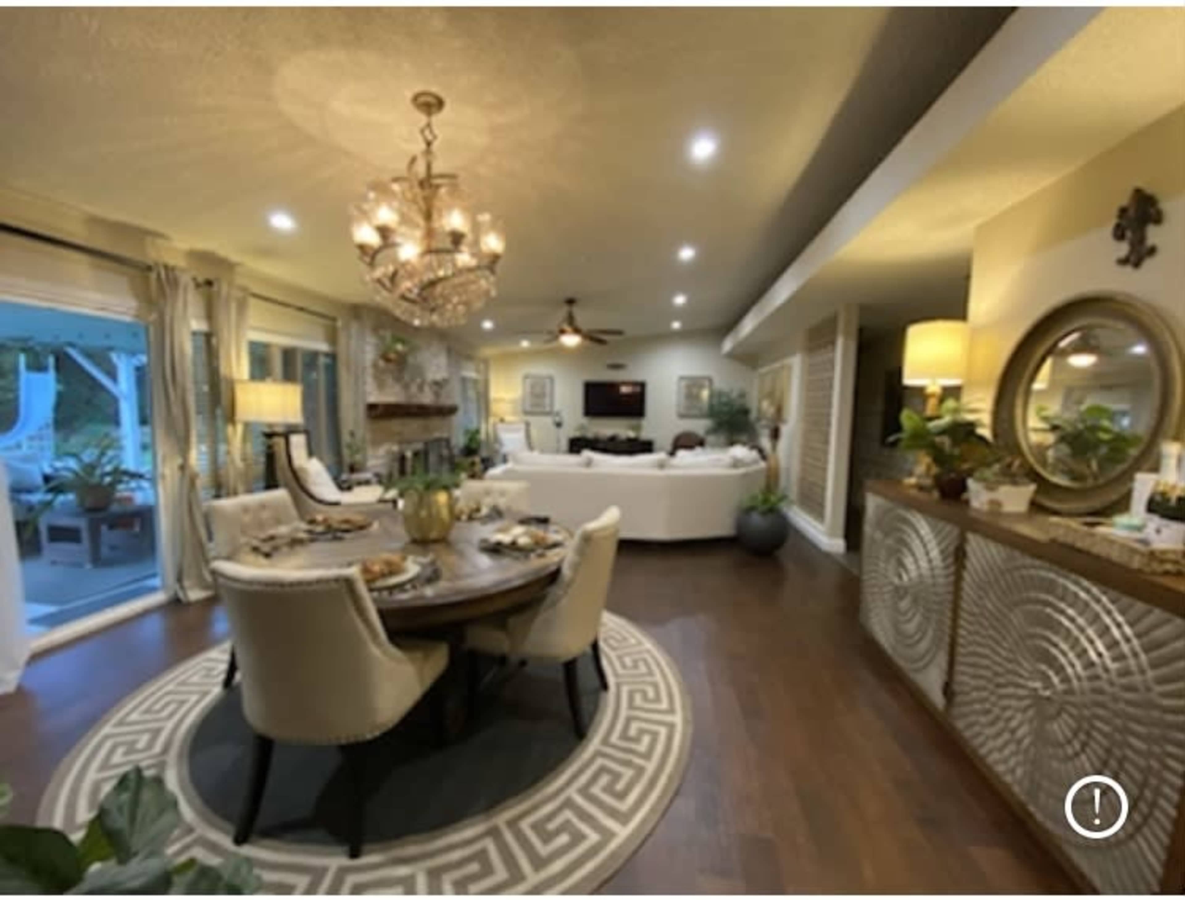 A well-decorated living and dining area featuring a round dining table set for a meal, a chandelier overhead, and a spacious seating arrangement in the background.