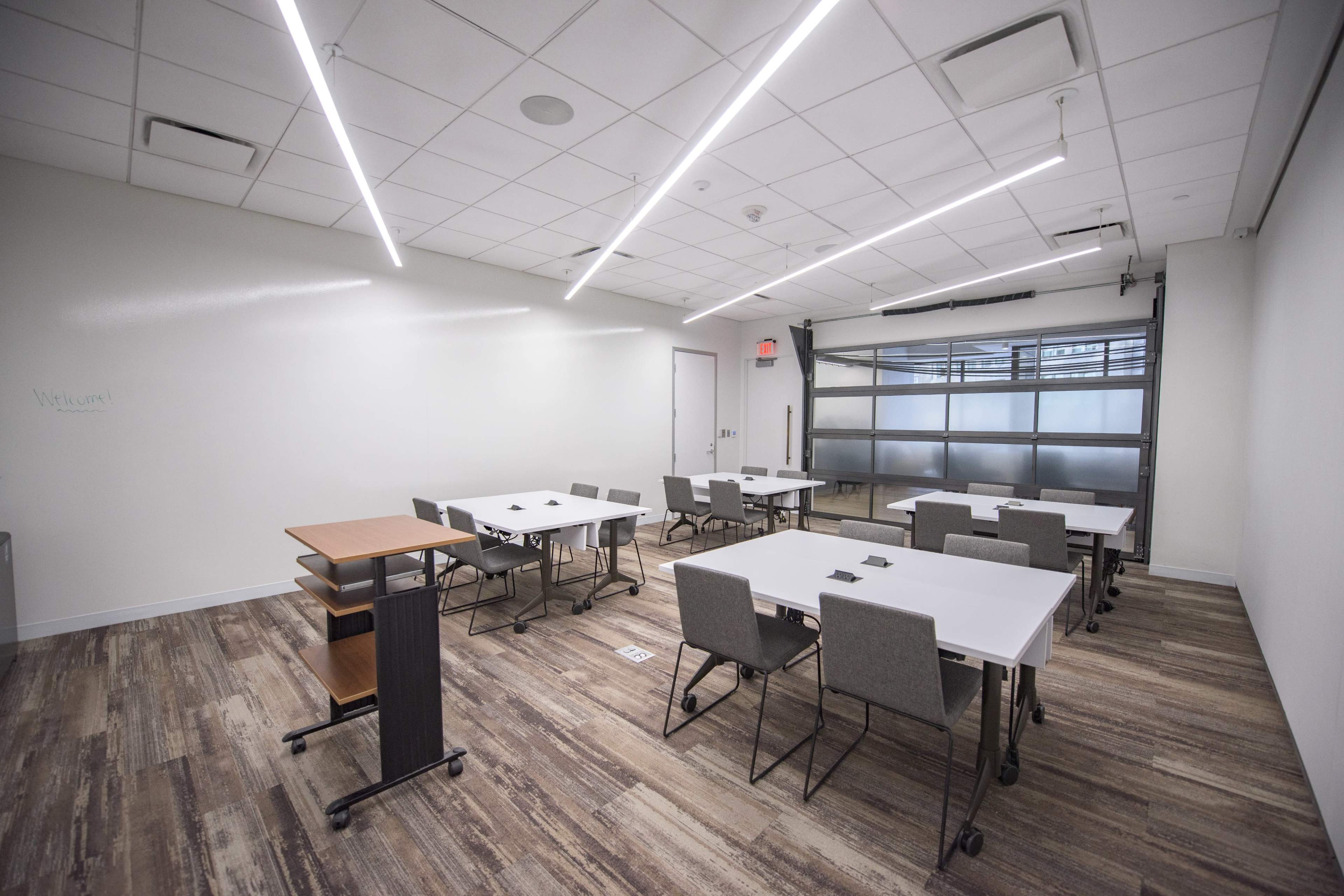 The image shows a modern classroom with multiple rectangular tables and chairs arranged in a spacious layout.