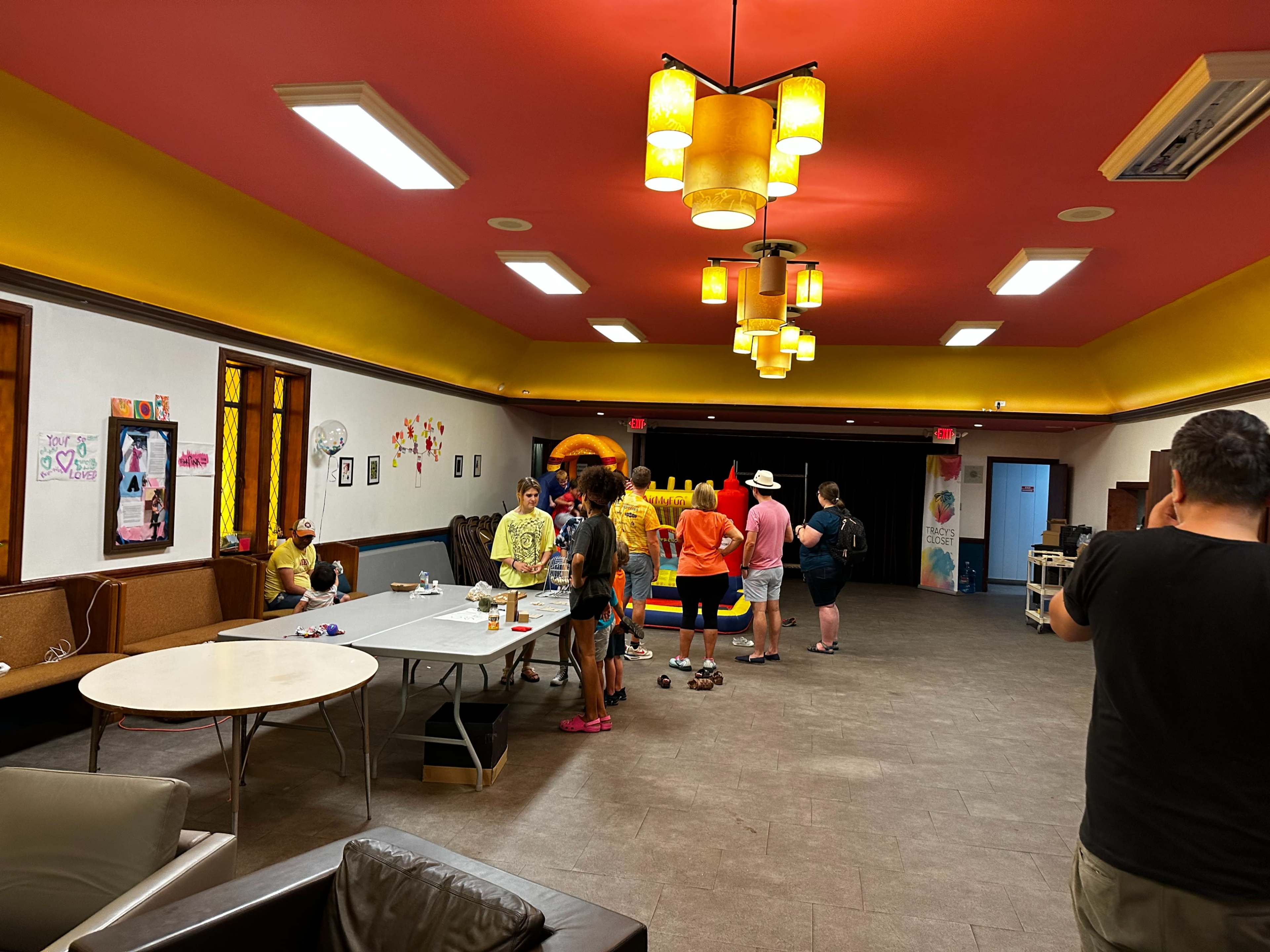 A group of people gathers in a brightly lit community room with yellow and orange decor, engaging around a table.
