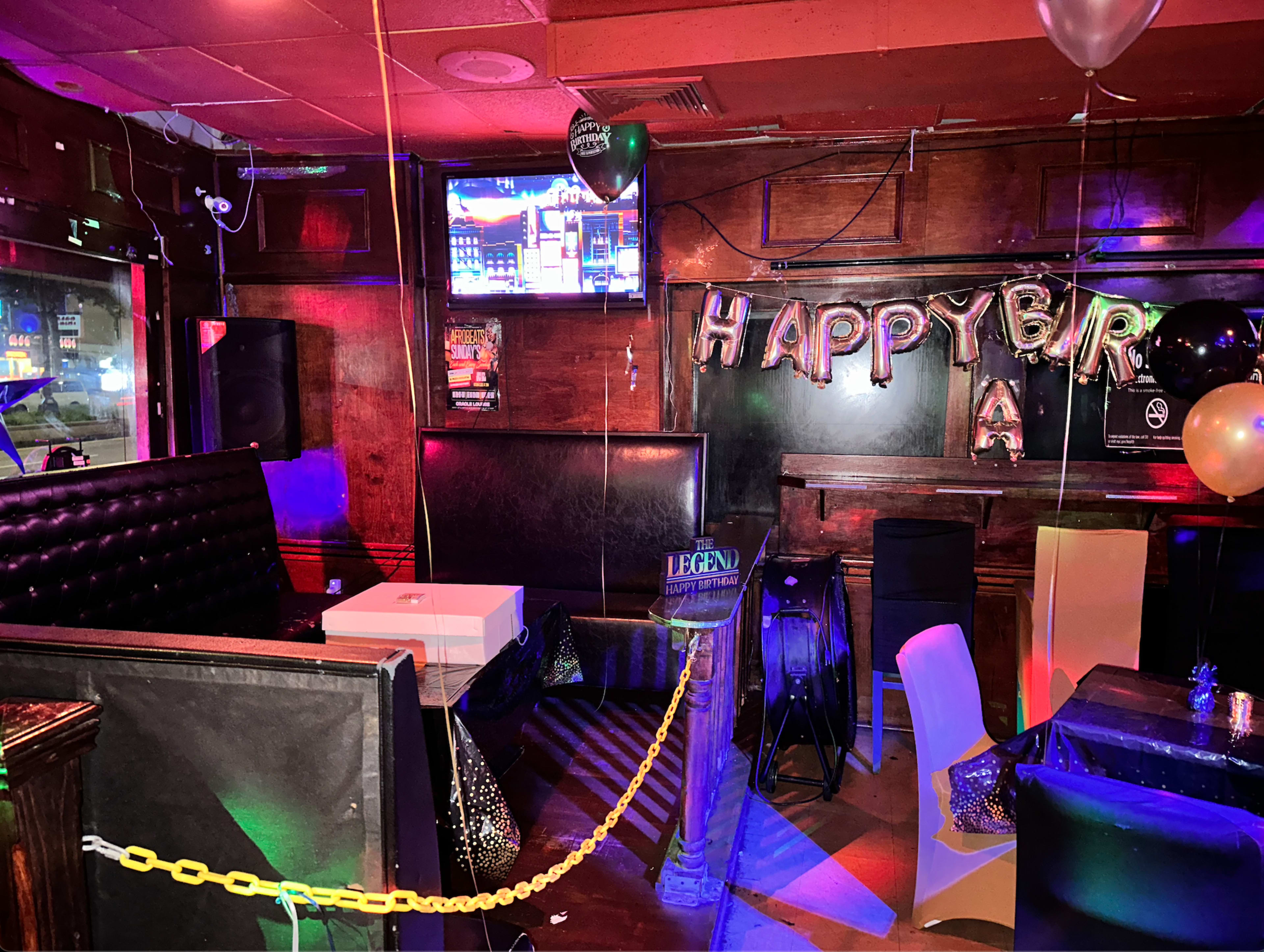 A dimly lit bar decorated with "Happy Birthday" balloons, a television screen, and several empty chairs and tables.