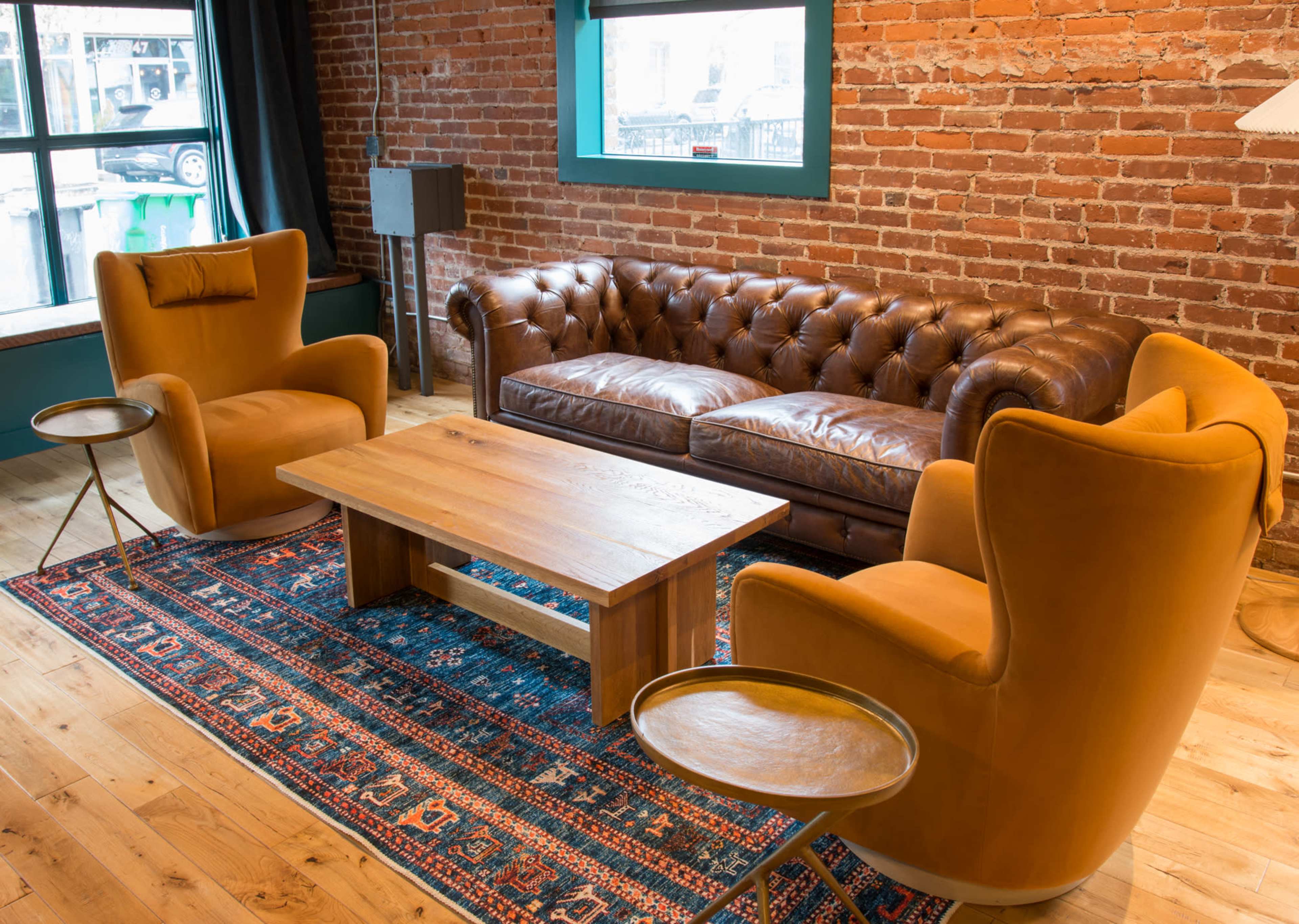 A cozy interior with a brown leather sofa, two yellow armchairs, a wooden coffee table, and a patterned rug on a wooden floor, set against a brick wall.