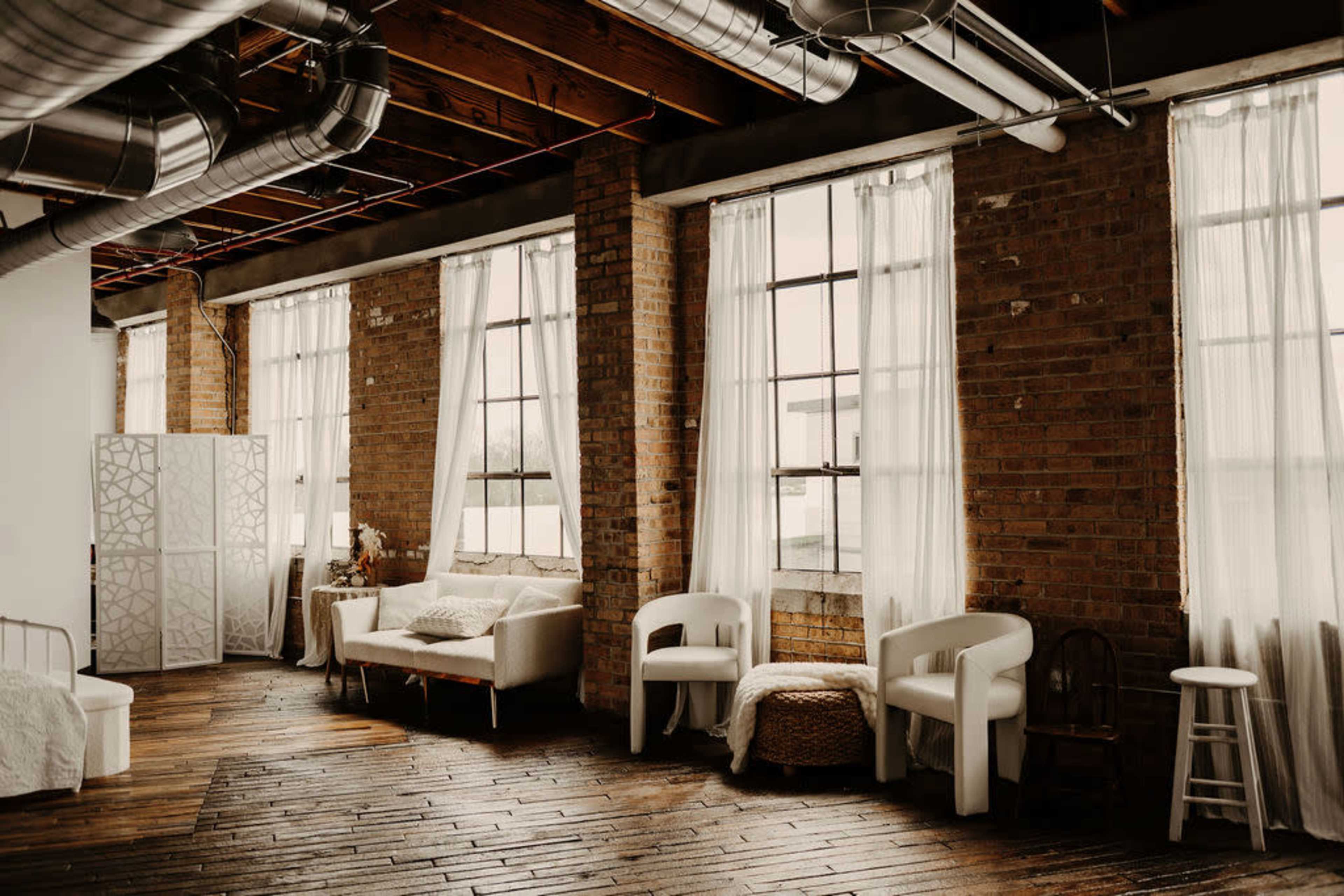 The image shows a spacious interior with large windows, exposed brick walls, and wooden flooring, furnished with a white sofa, chairs, and a decorative screen.