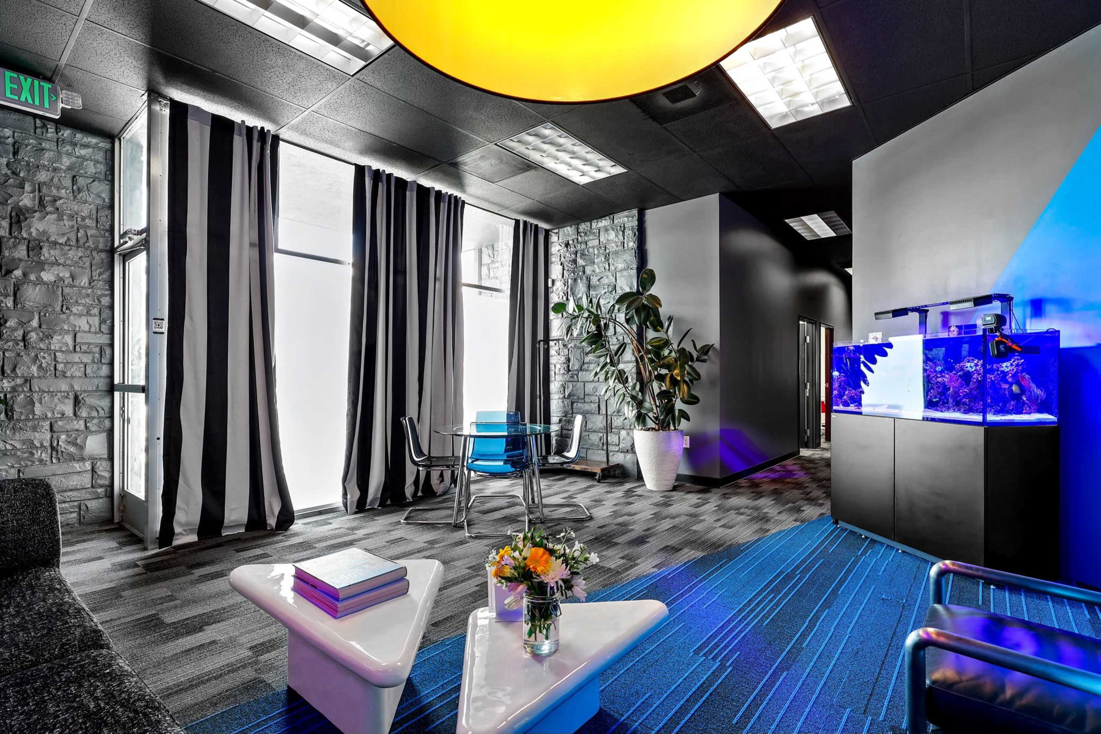 The image shows a modern lounge area featuring a black and white striped curtain, a transparent dining table with chairs, an aquarium, and a geometric coffee table with a floral arrangement.