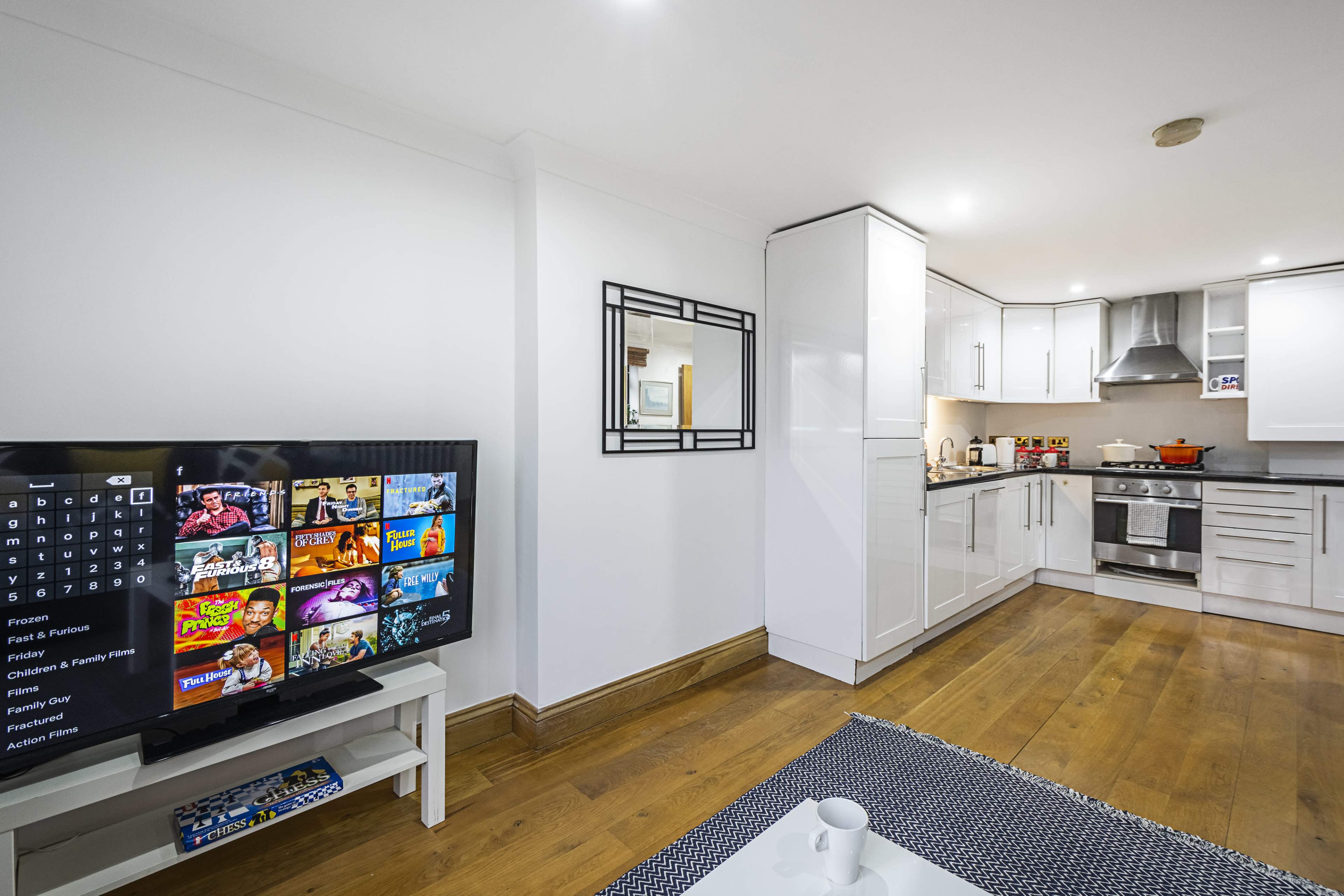 The image shows a modern, open-concept living area featuring a television on a stand, a mirror on the wall, and a kitchen with white cabinets and stainless steel appliances.