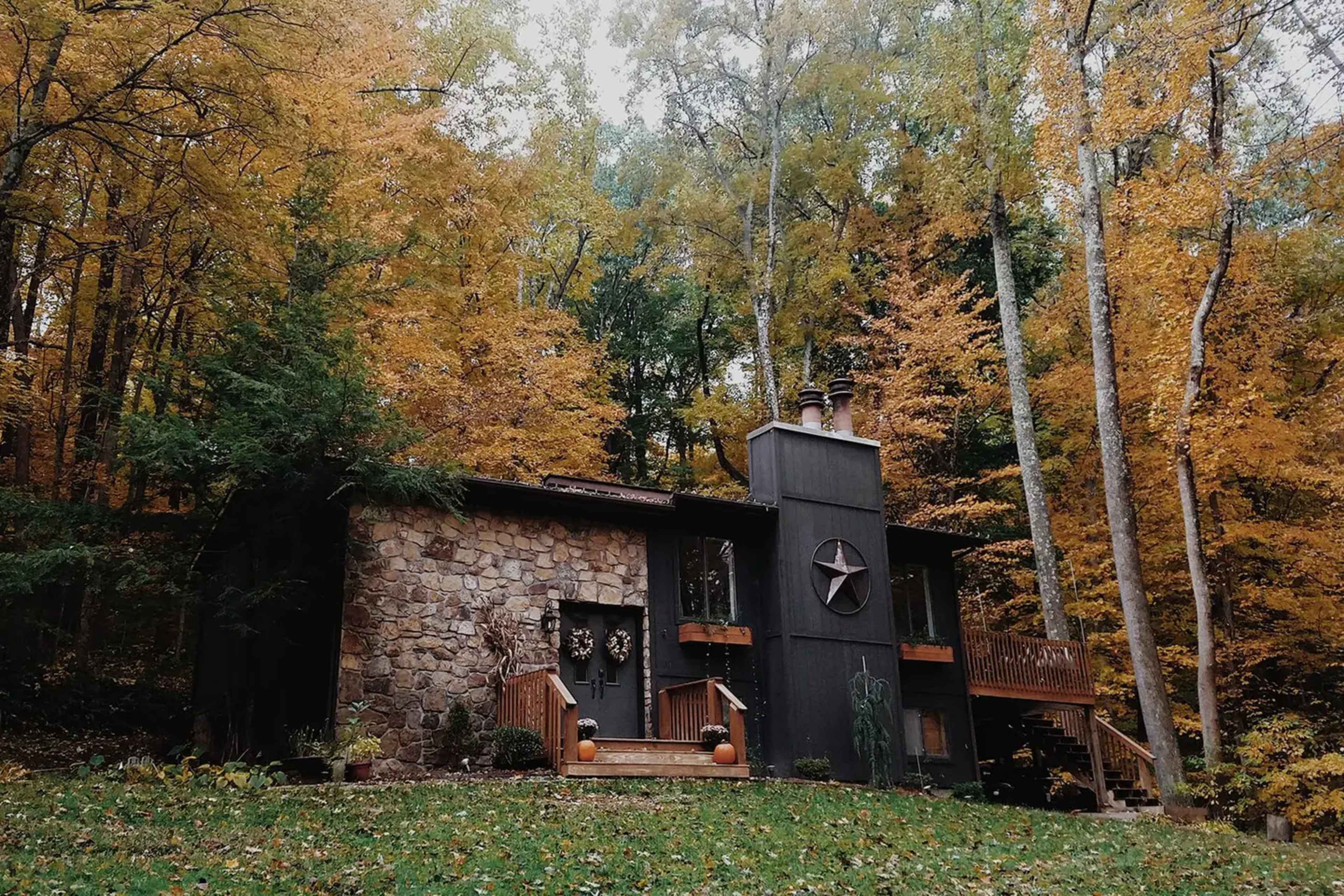 A stone and wooden cabin is surrounded by trees with autumn foliage, featuring a porch and a star decoration on the front.