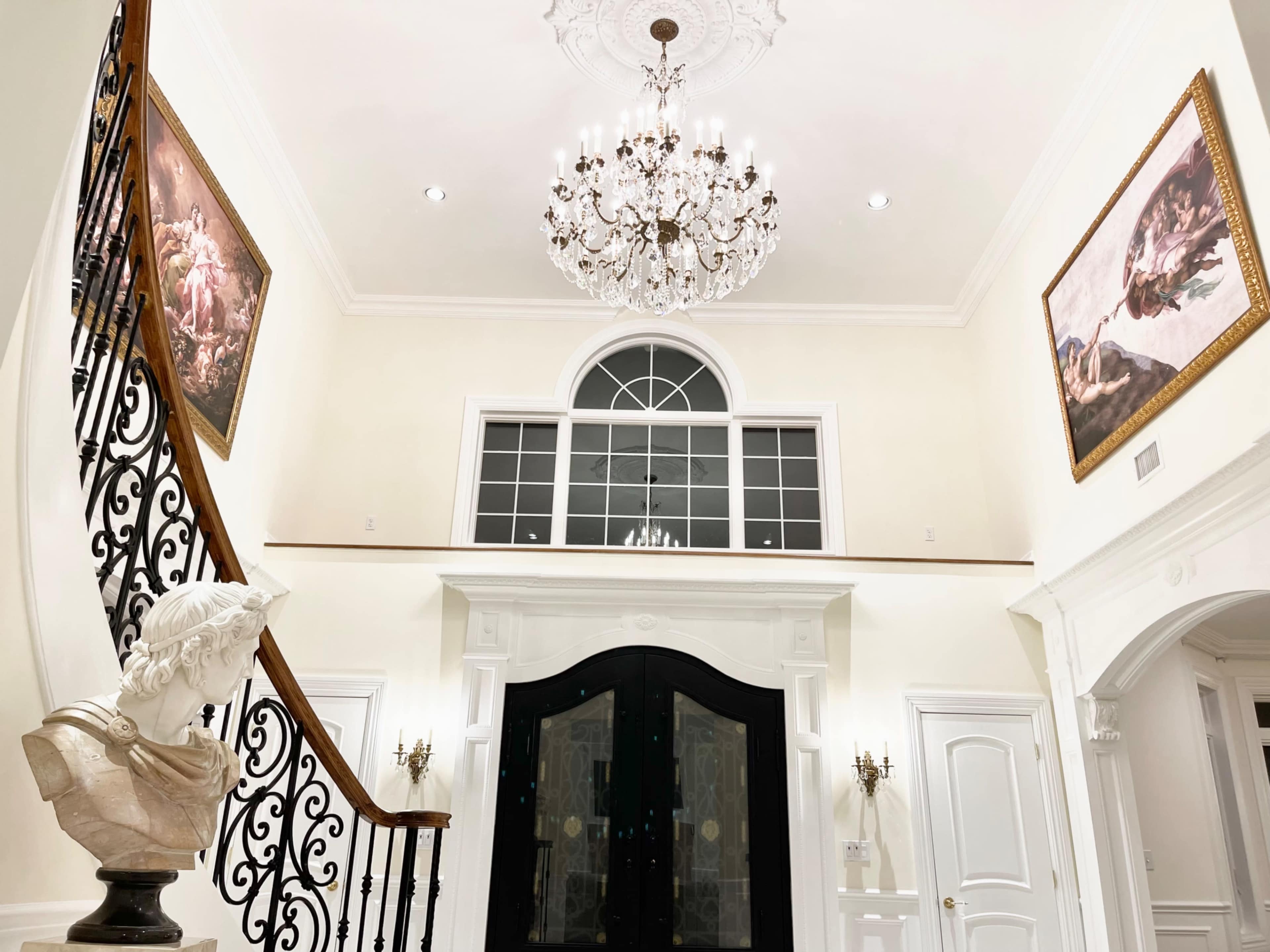 A grand foyer features a large chandelier, an ornate staircase with a wrought iron railing, and classical paintings on the walls.