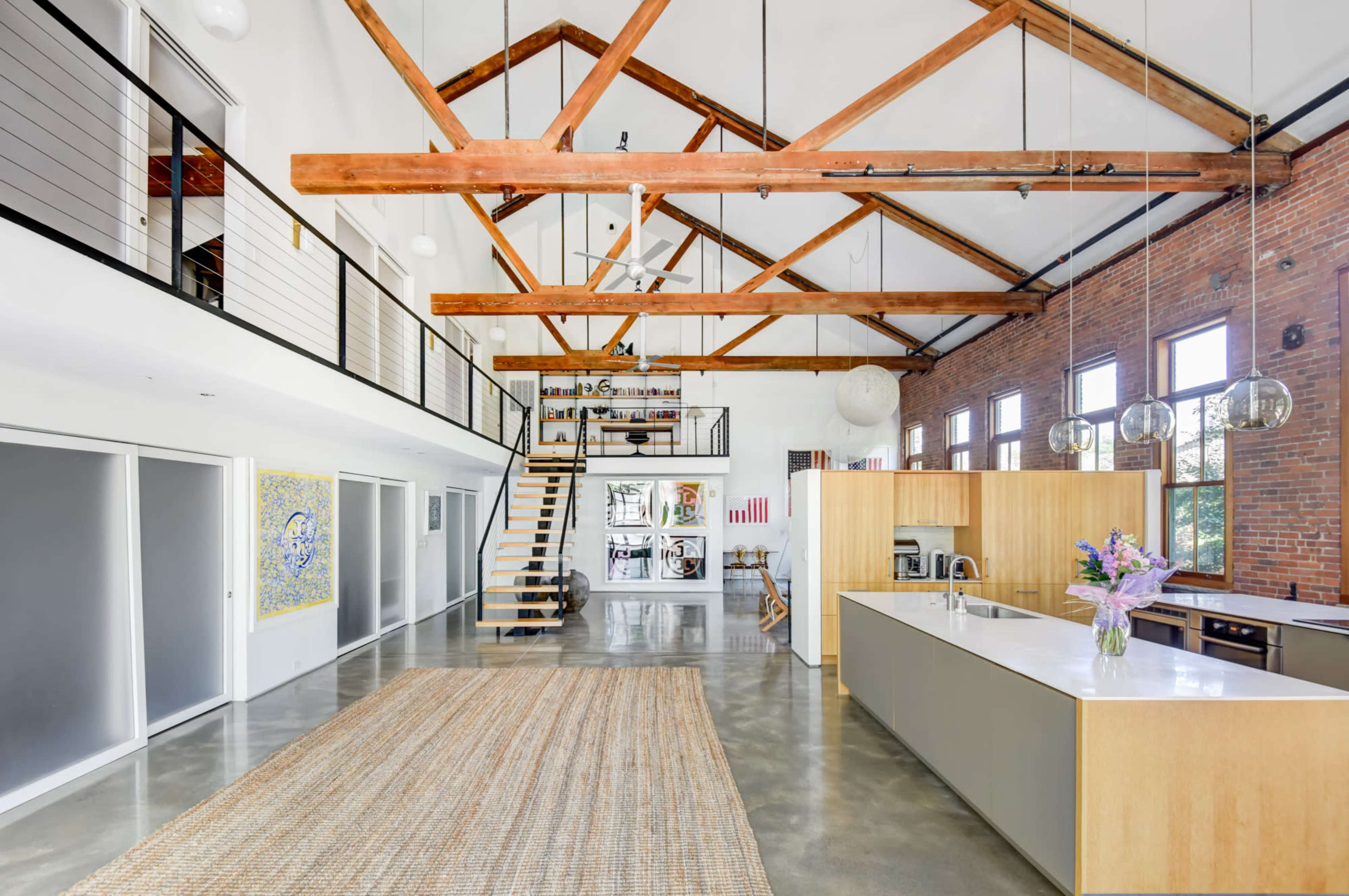 An open-concept loft with high ceilings, exposed wooden beams, and a central kitchen island, complemented by a staircase leading to an upper level.