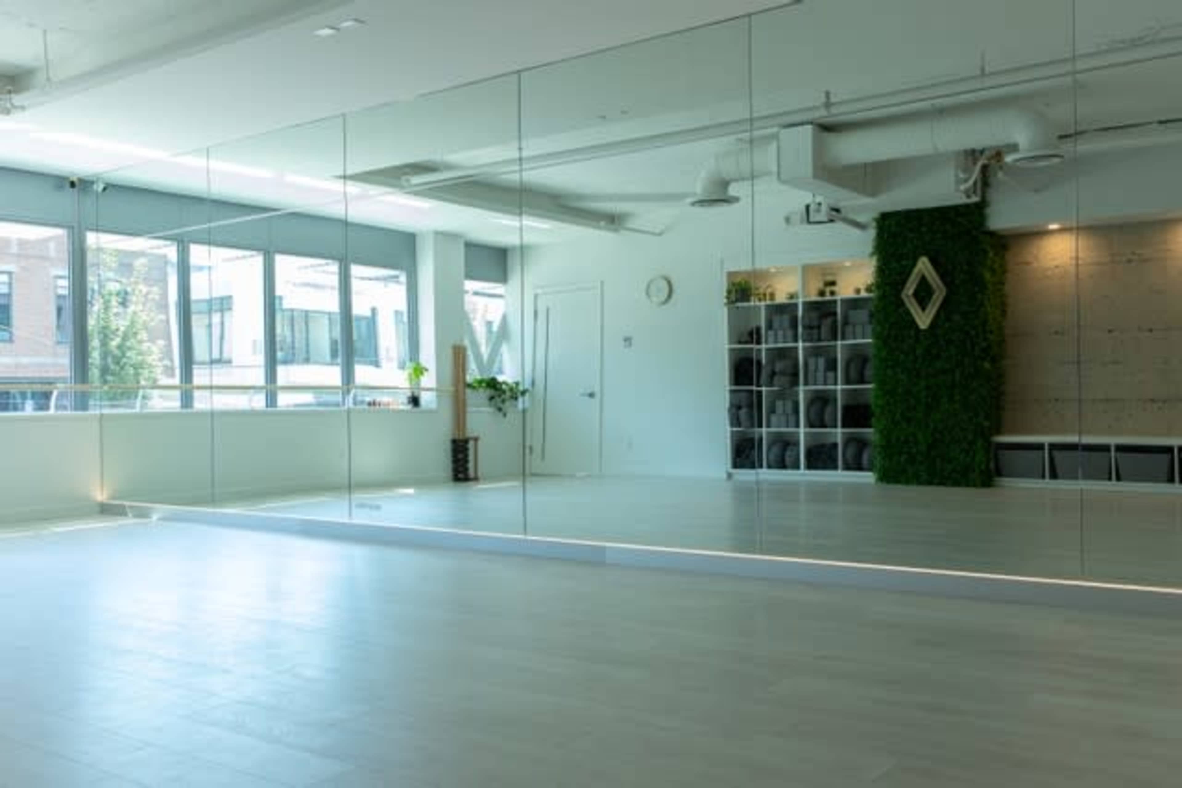 A bright, minimalist studio features large mirrors on one wall, a green plant, and a shelf with neatly arranged items.