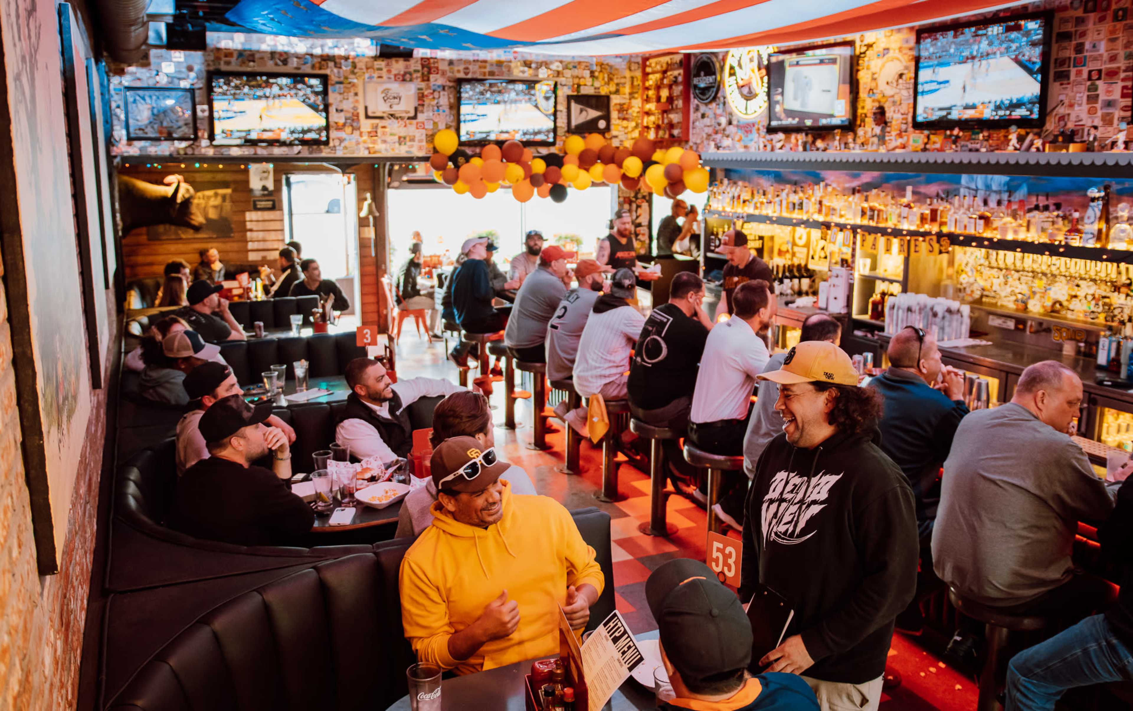 The image shows a lively bar scene filled with patrons sitting at tables and the bar, with multiple televisions displaying a sports event and colorful decorations in the background.