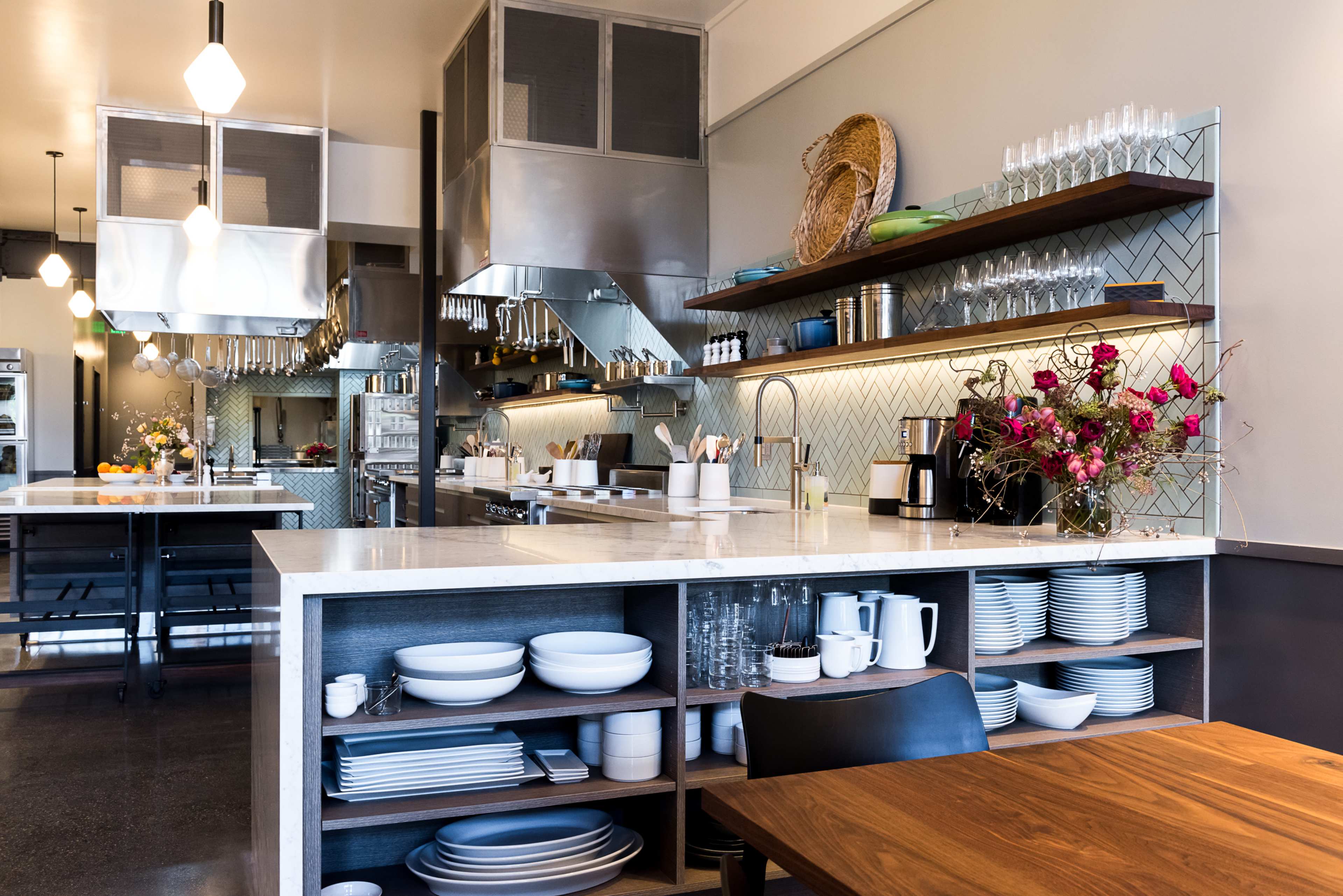A modern kitchen features stainless steel appliances, open shelves with glassware, and a large island with white dishes and a wooden table.