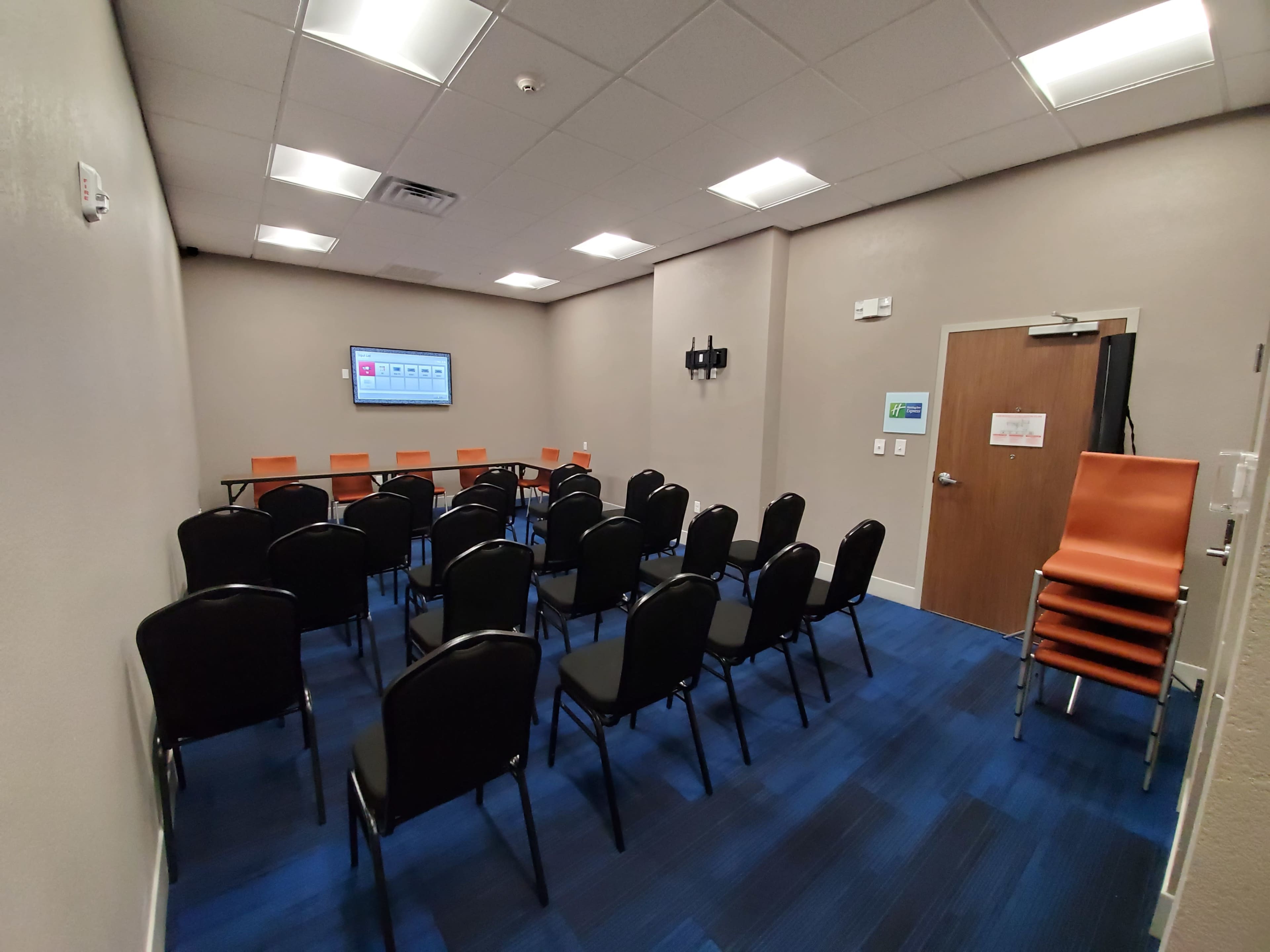 A small meeting room with a row of black chairs facing a screen and a table, with additional stacked orange chairs in the corner.