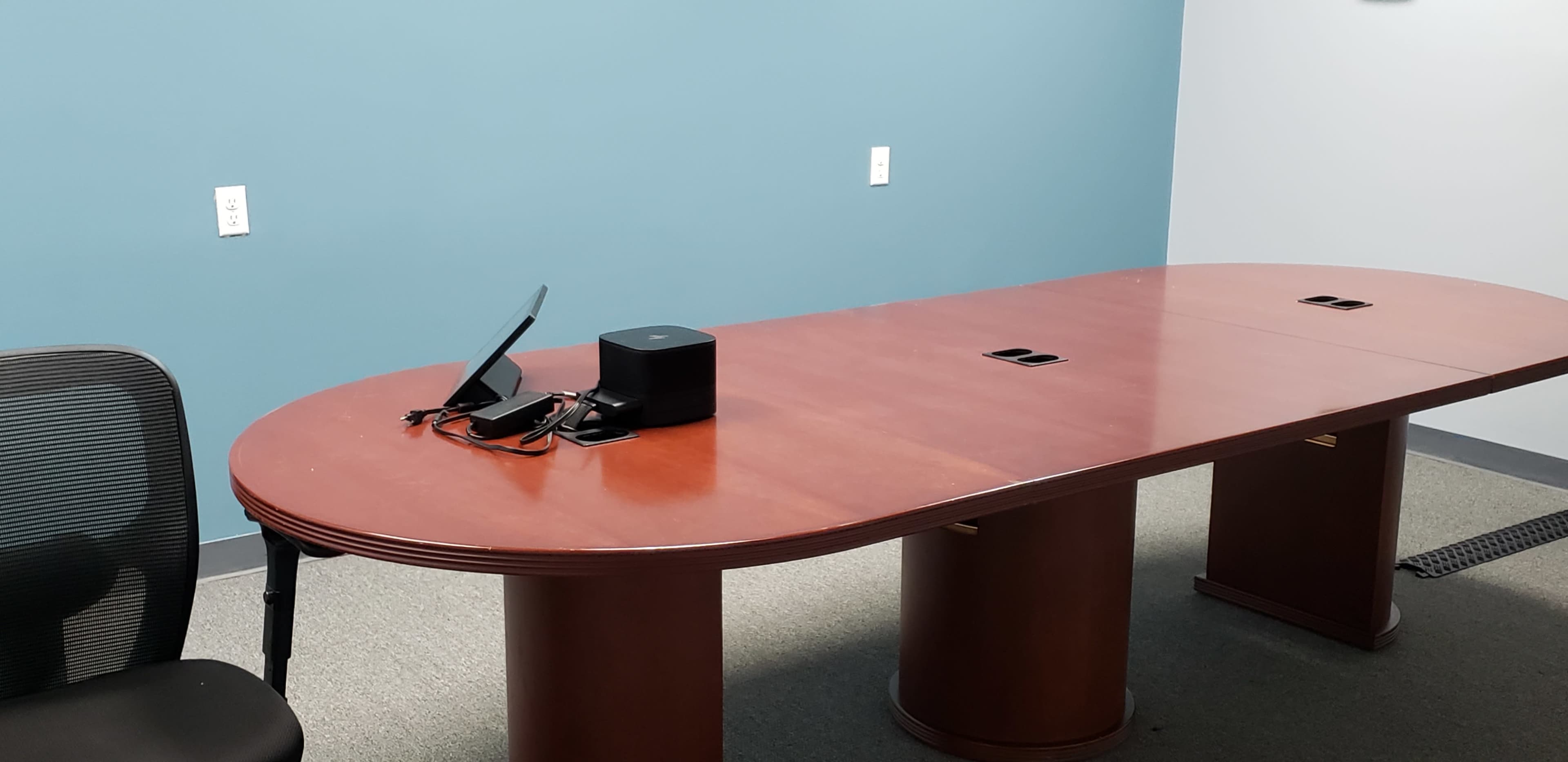 A large wooden conference table is positioned in a room with blue walls, accompanied by a black chair and a small electronic device on the table.