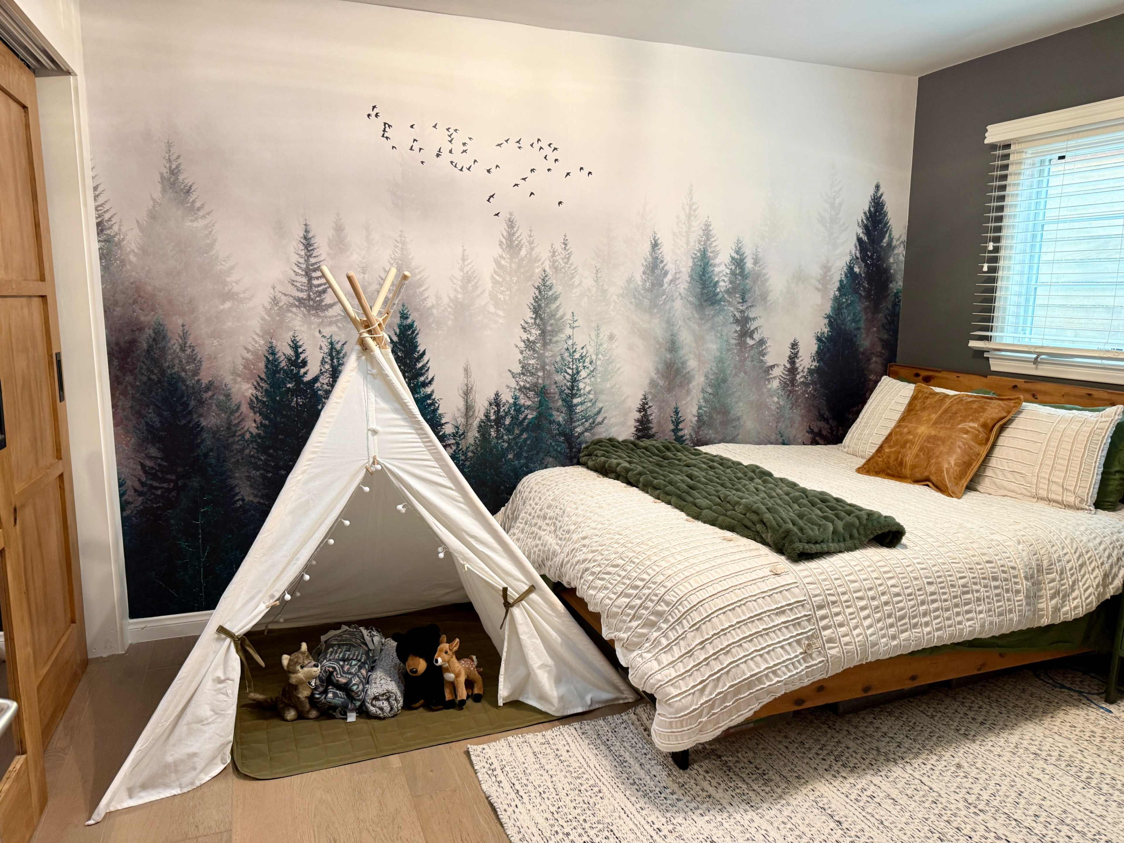 The image features a cozy bedroom with a forest-themed mural on the wall, a bed with textured bedding, and a children's play tent filled with plush toys.