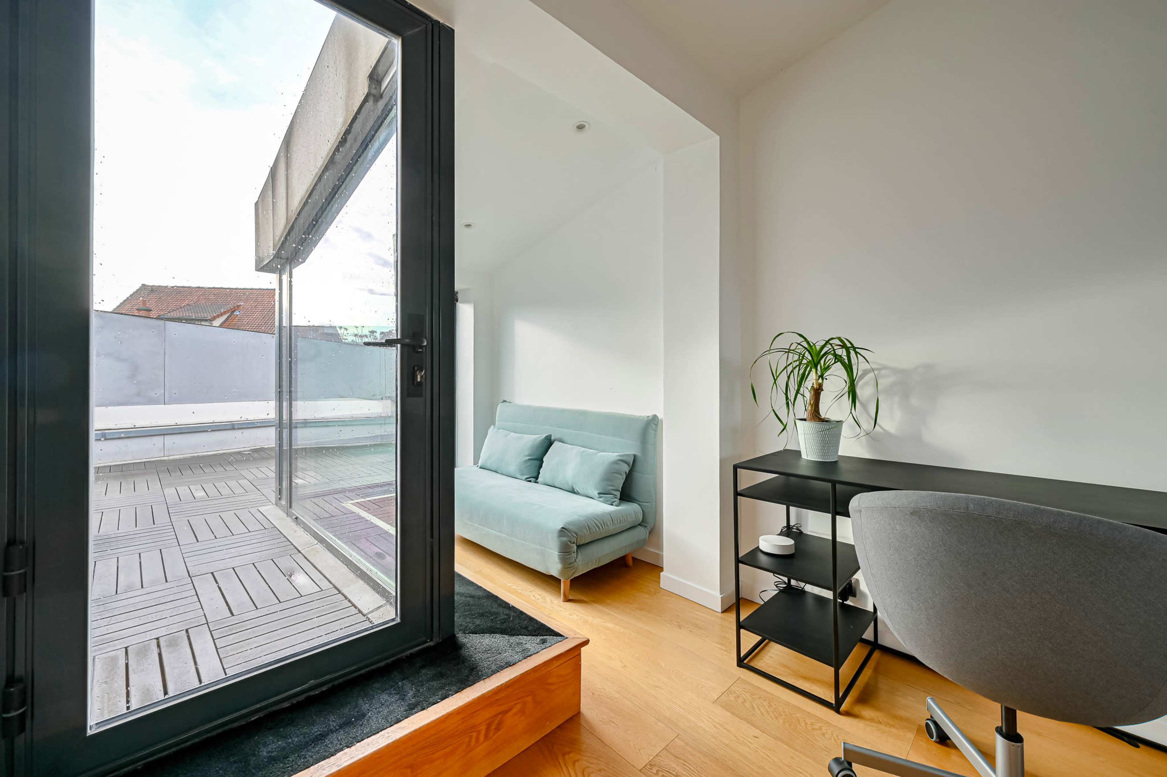A small, modern room features a sliding glass door opening onto a terrace, a light blue sofa, and a minimalist black desk with a plant.