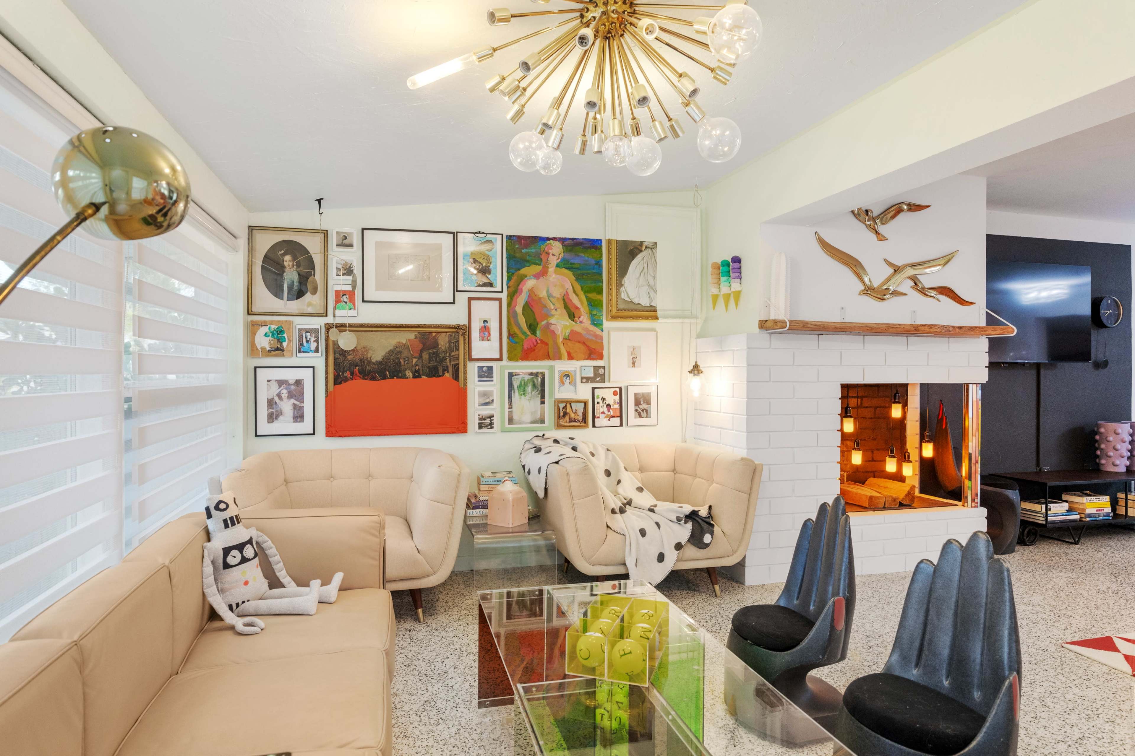 A modern living room features two beige sofas, a glass coffee table, various artworks on the walls, and a fireplace with decorative elements.