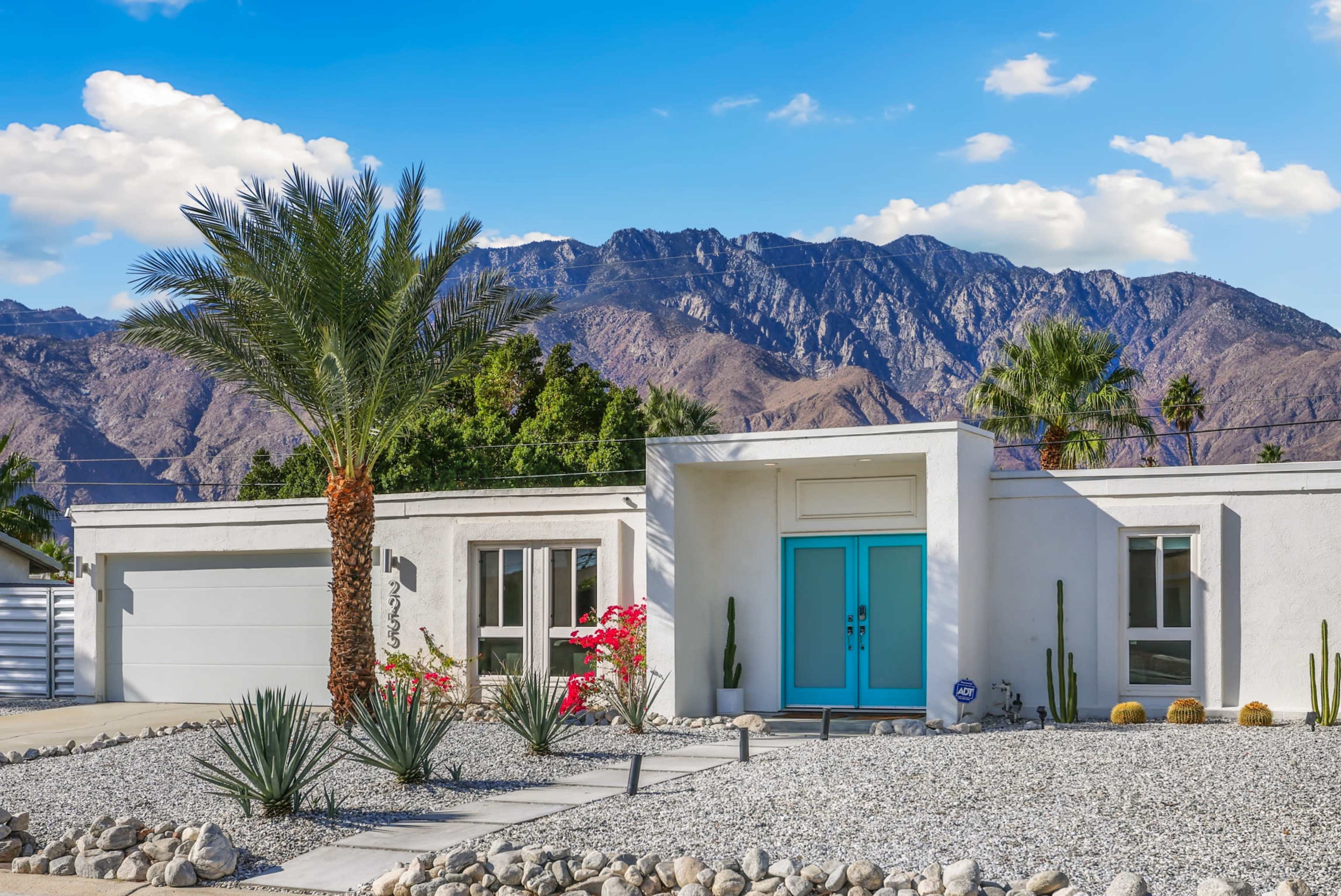 A modern single-story house with a turquoise door is surrounded by desert landscaping and mountains in the background.