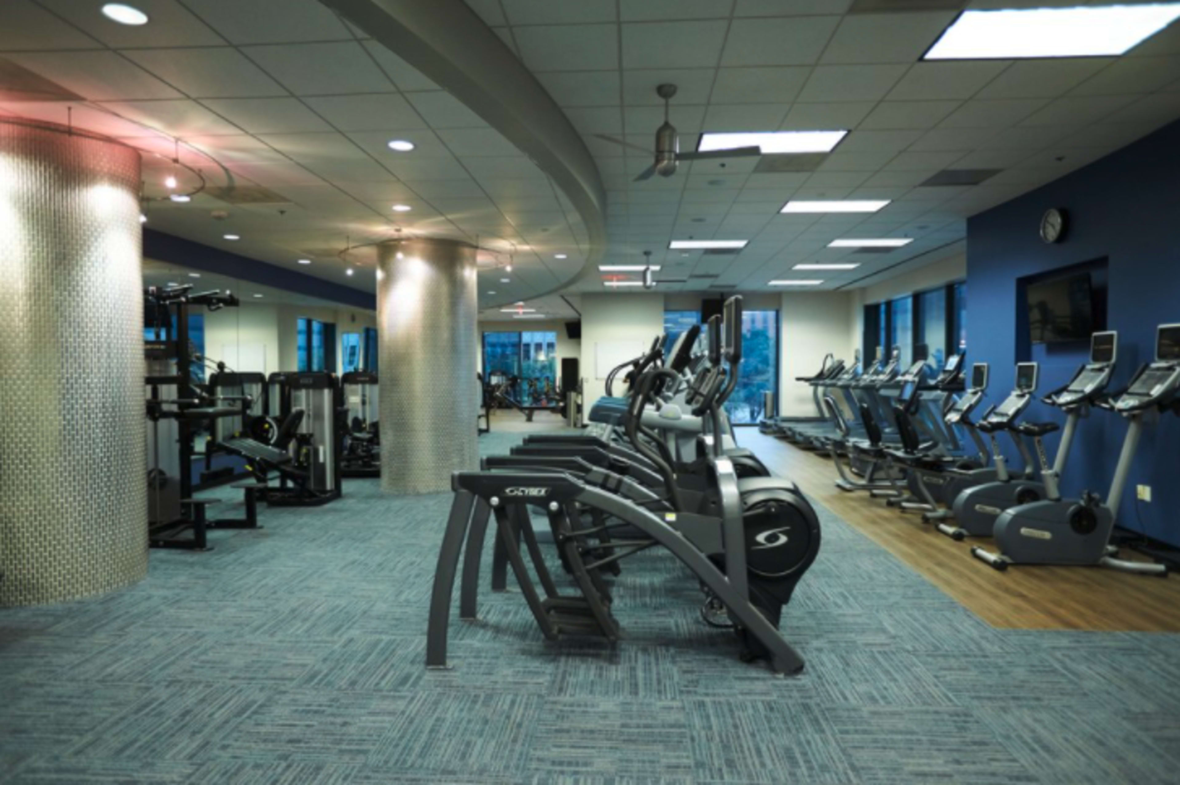 The image shows a modern gym interior with various exercise machines and equipment arranged in an open, well-lit space.