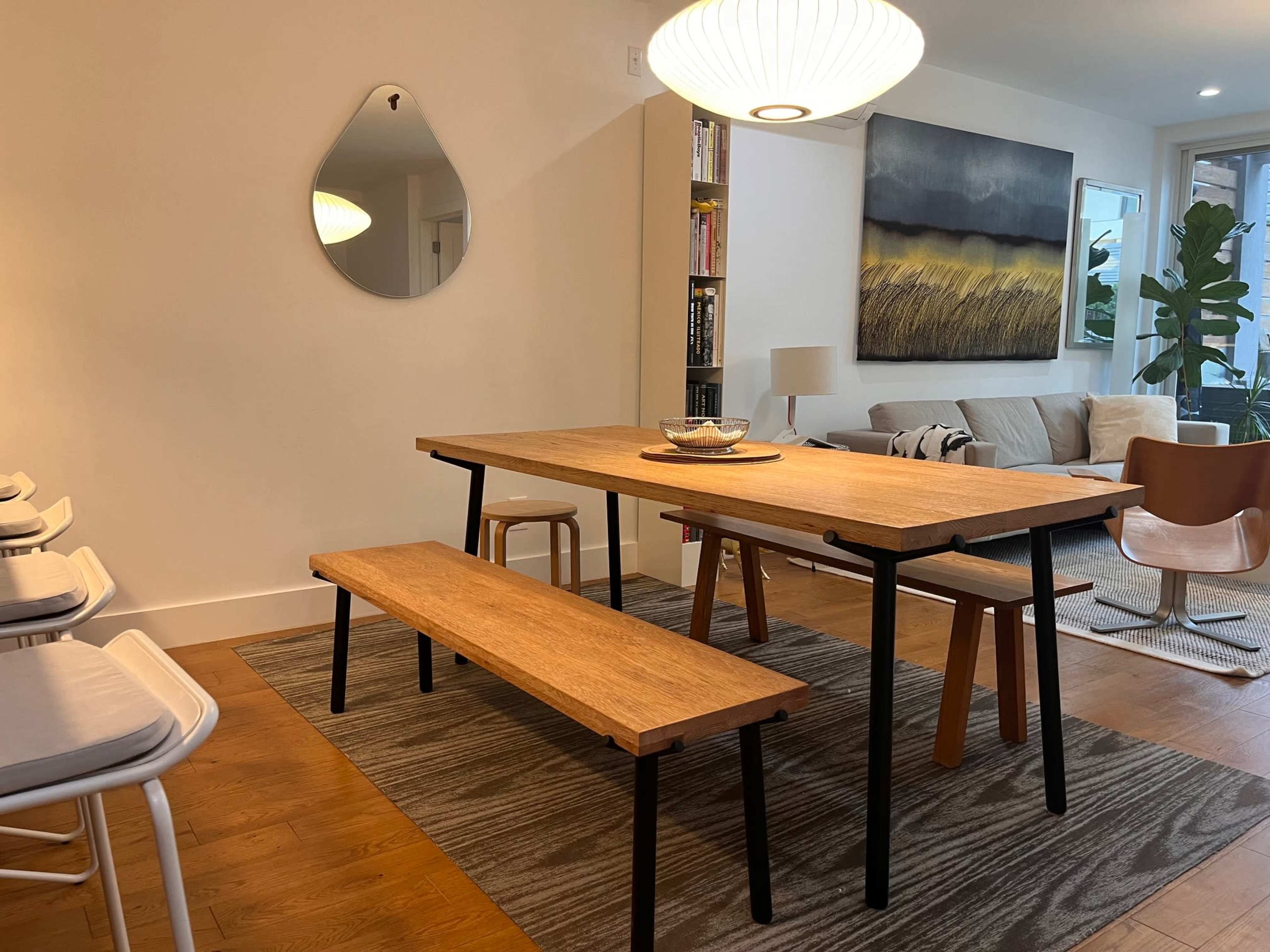 A wooden dining table with a bench and chairs is positioned in a modern living area that features a large painting and a plant.