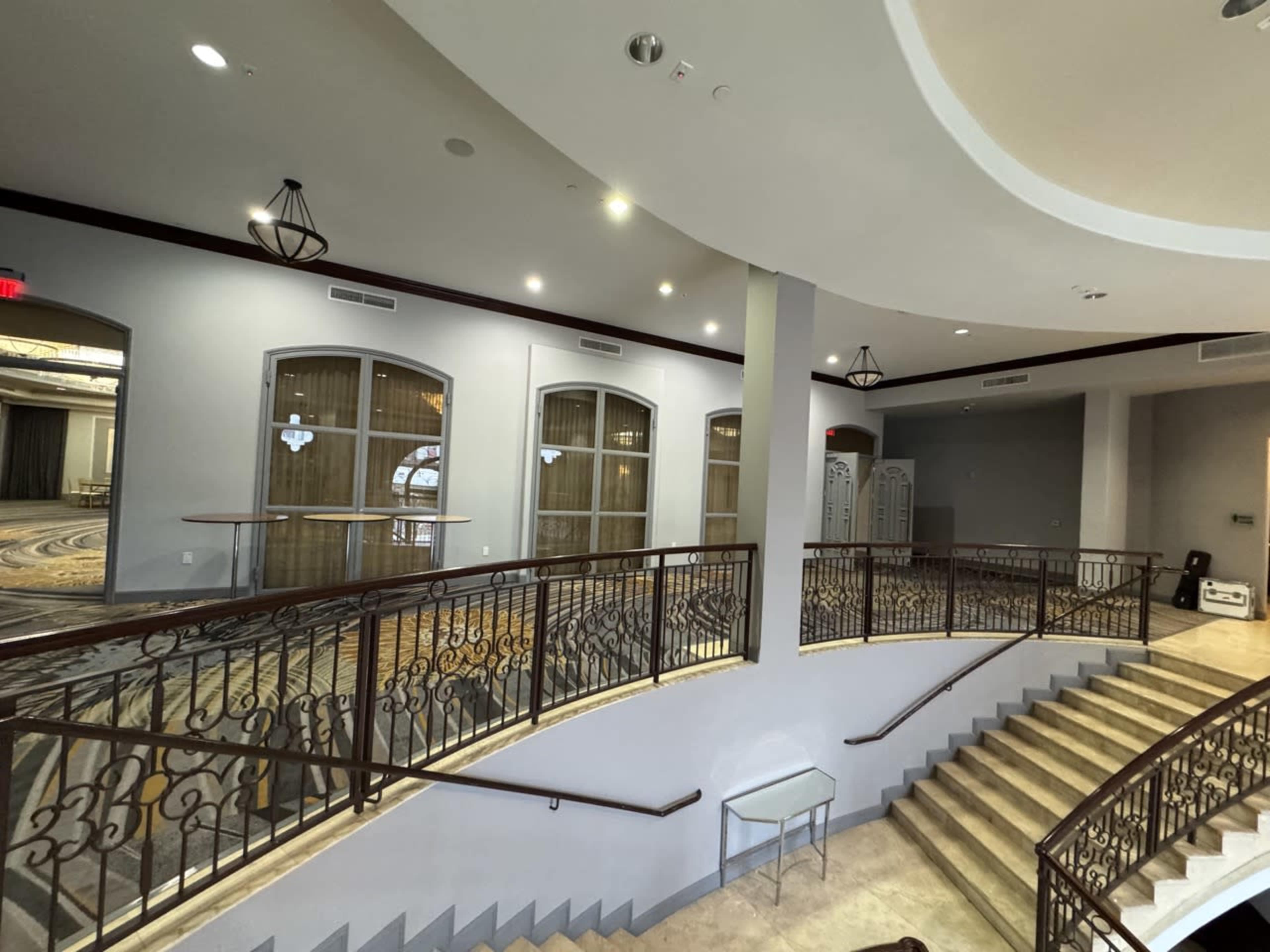 The image shows a modern interior space featuring a staircase with iron railing, illuminated by overhead lights, and a large, open area with light-colored walls and windows.