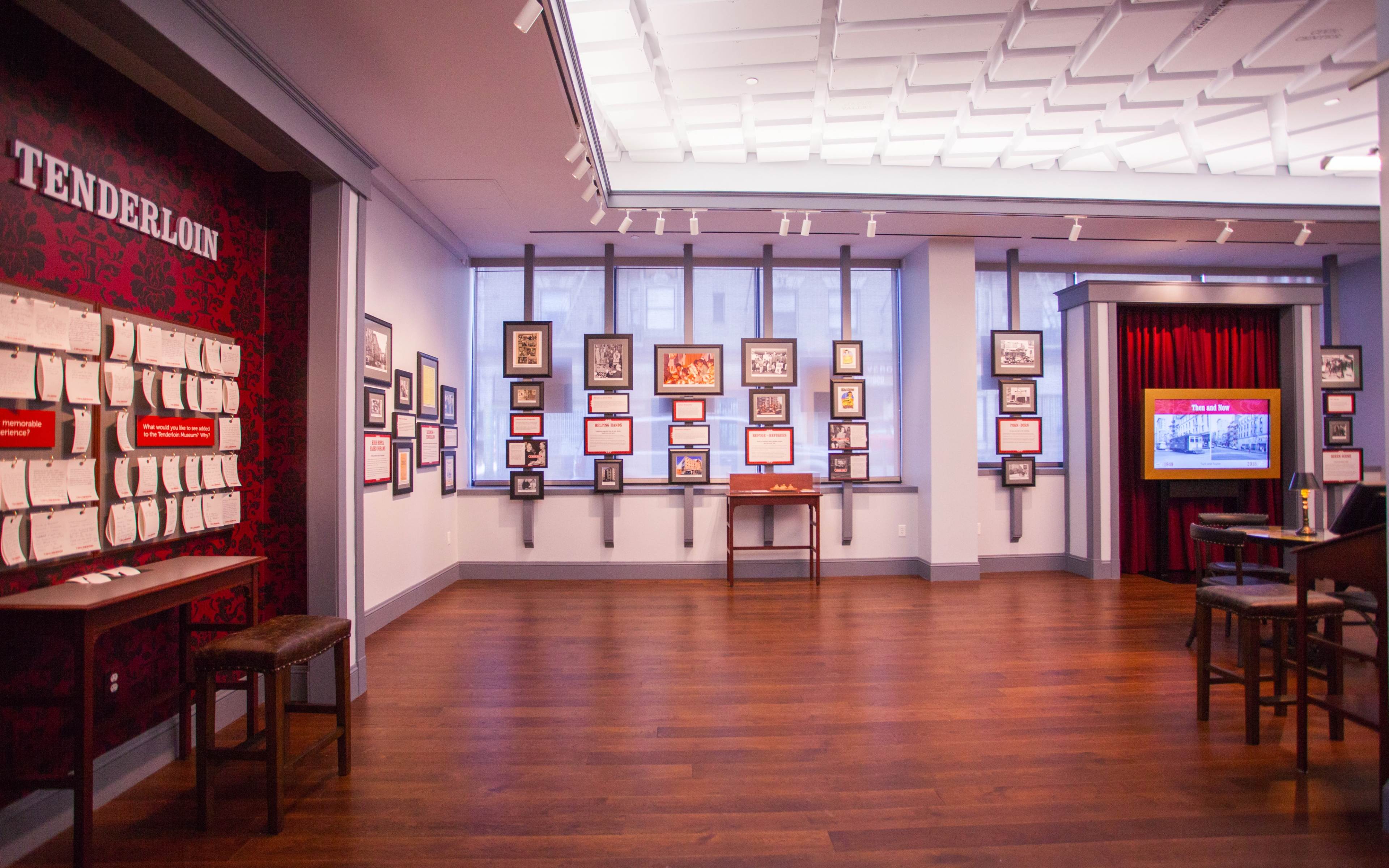 A museum exhibit featuring a series of framed photographs and display boards on the walls, with a red velvet curtain and a video screen in one corner.