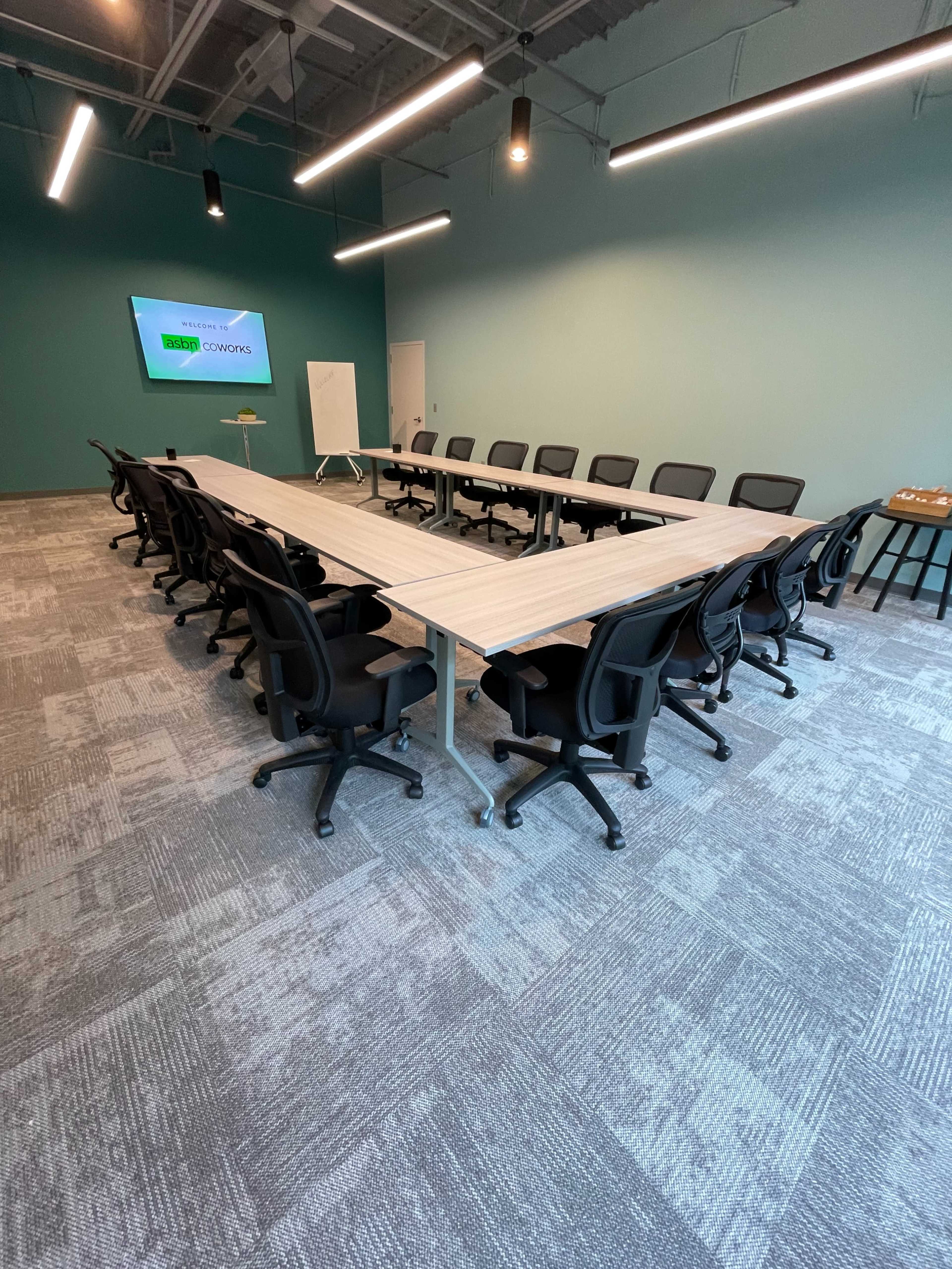 A rectangular conference room features a long table surrounded by black office chairs, with a large screen mounted on a light blue wall.