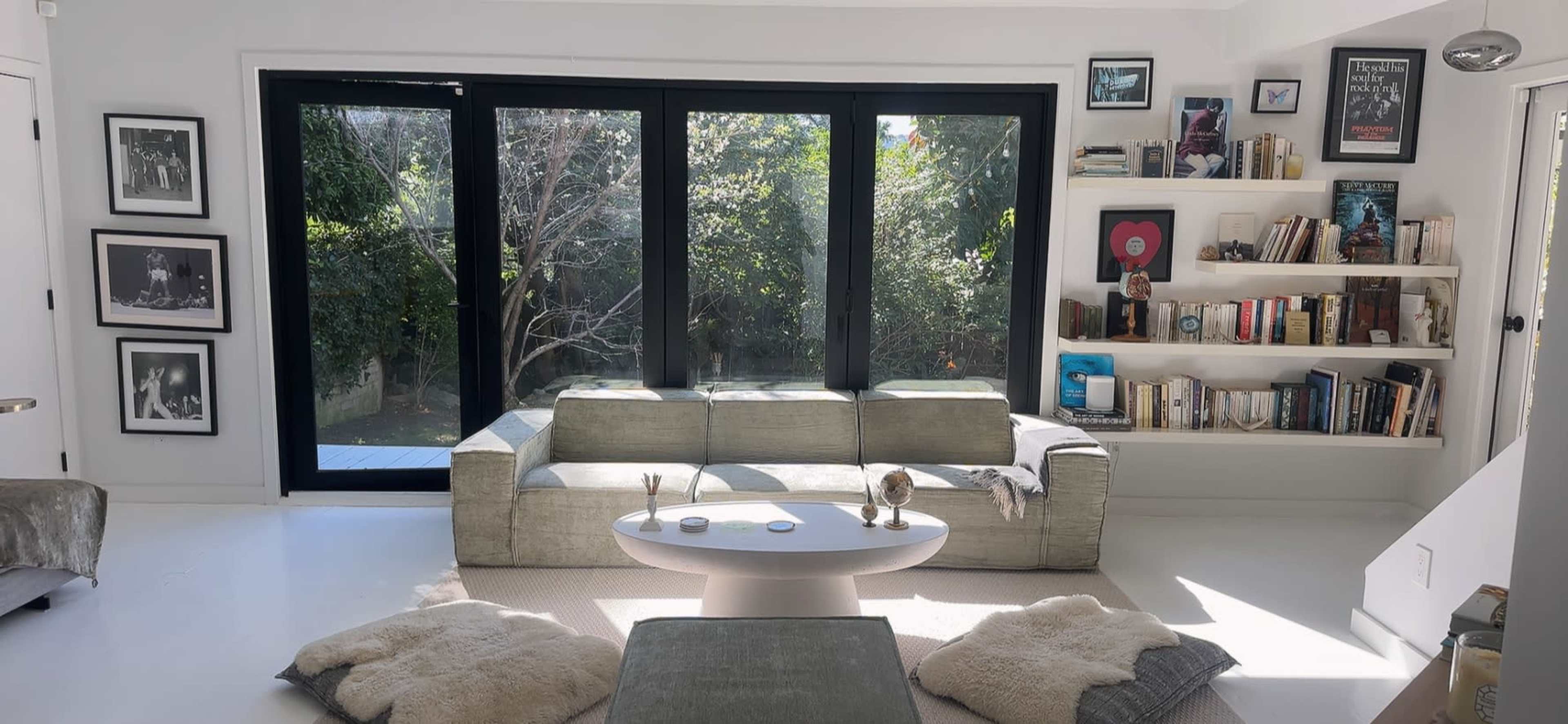 The image shows a modern living room featuring a gray couch, a circular table, and large windows overlooking a garden, with bookshelves and framed photos on the walls.