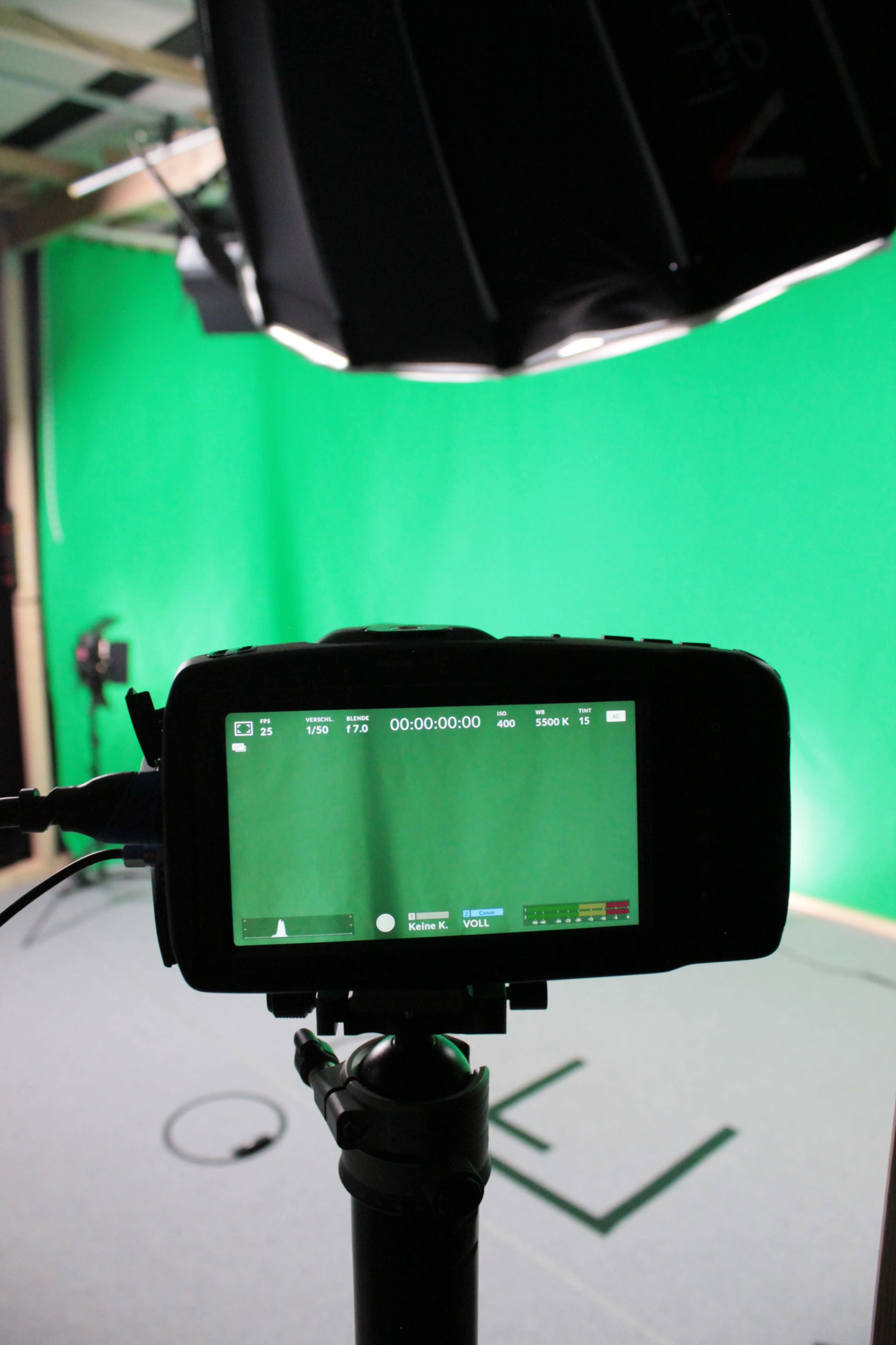 The image shows a camera monitor on a tripod, displaying a recording timer against a green backdrop in a studio setting.