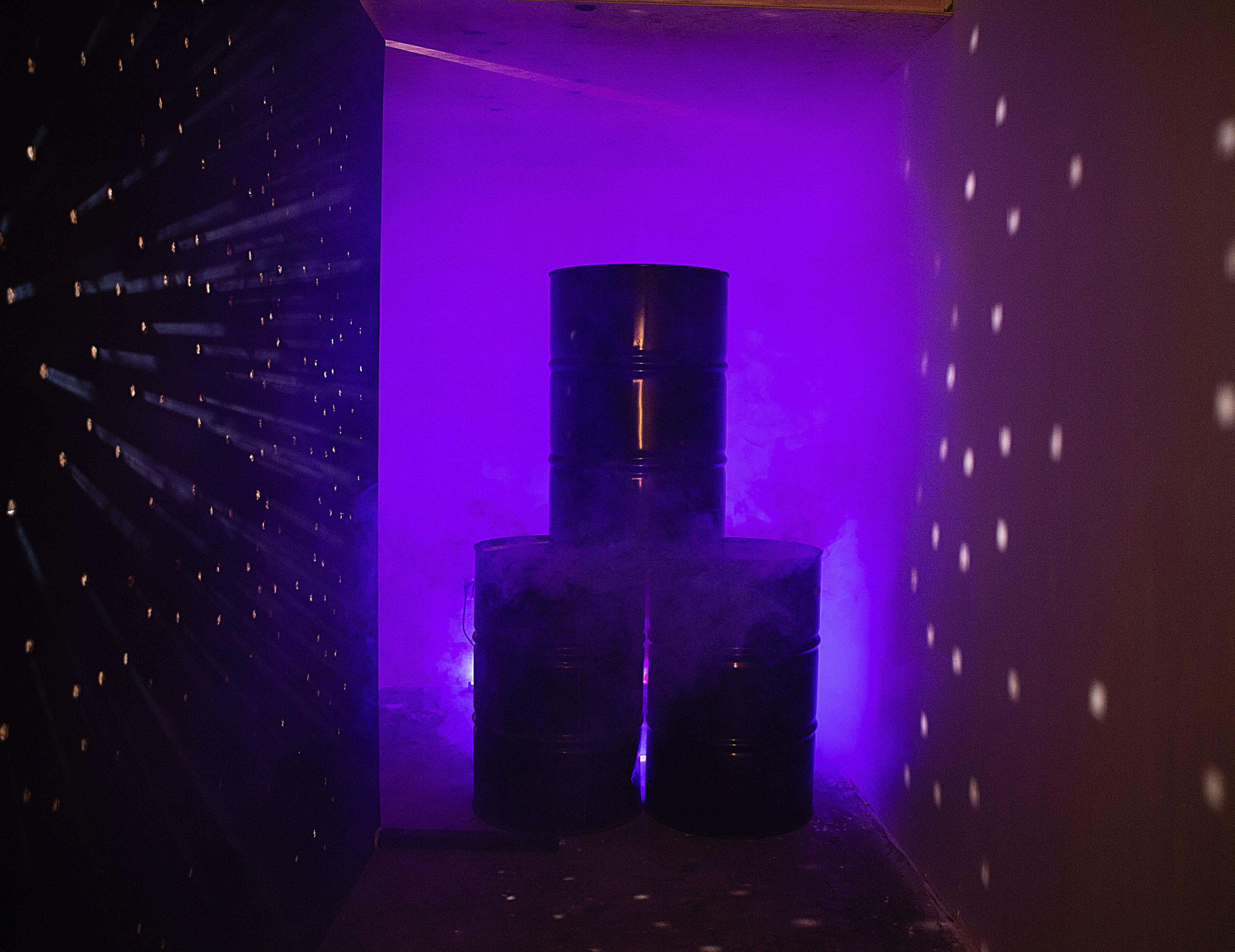 Three stacked black barrels surrounded by purple smoke and beams of light in a dimly lit corridor.