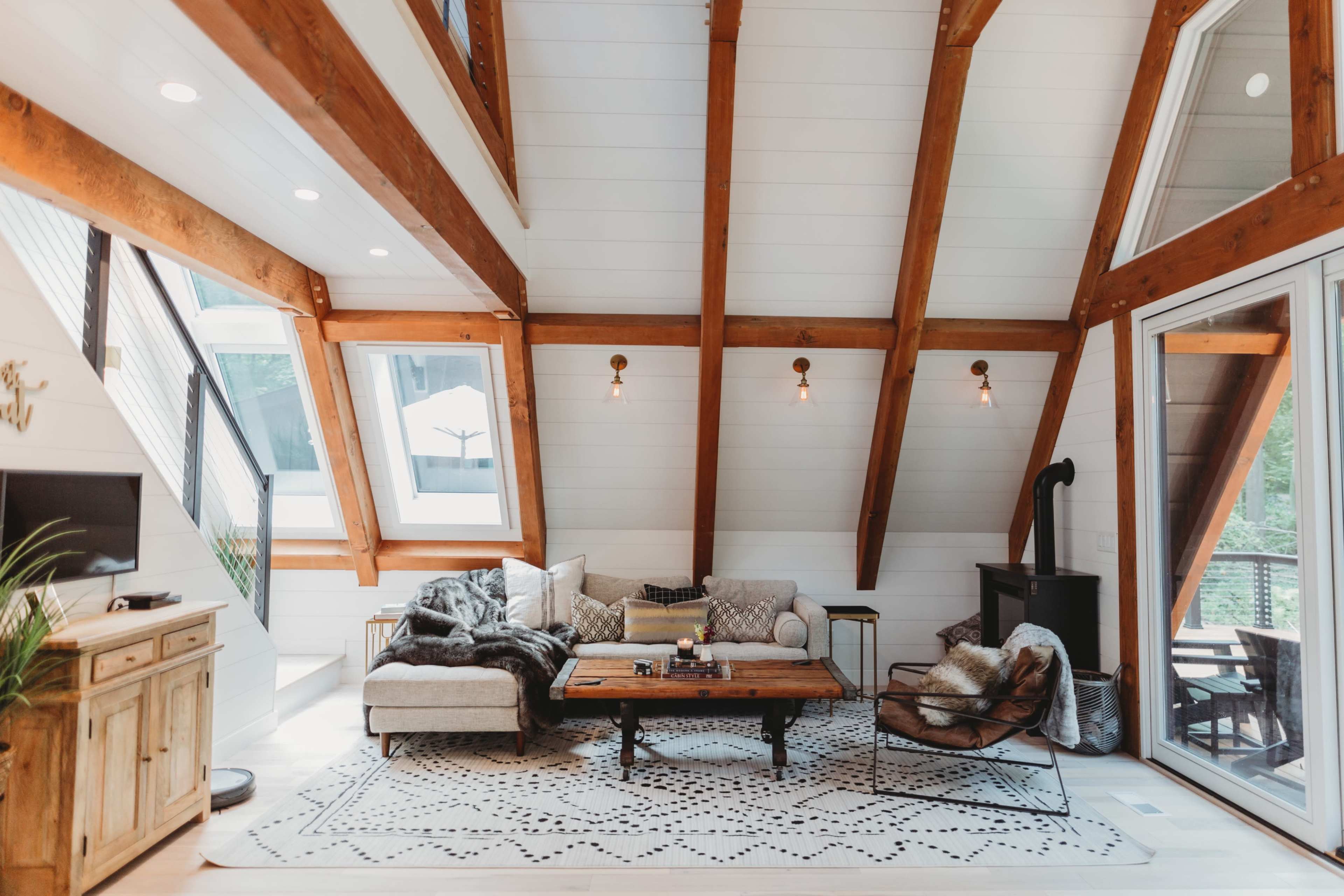 The image shows a modern living room with high, wooden beam ceilings, large windows, and a cozy arrangement of furniture including a sectional sofa and a wooden coffee table.