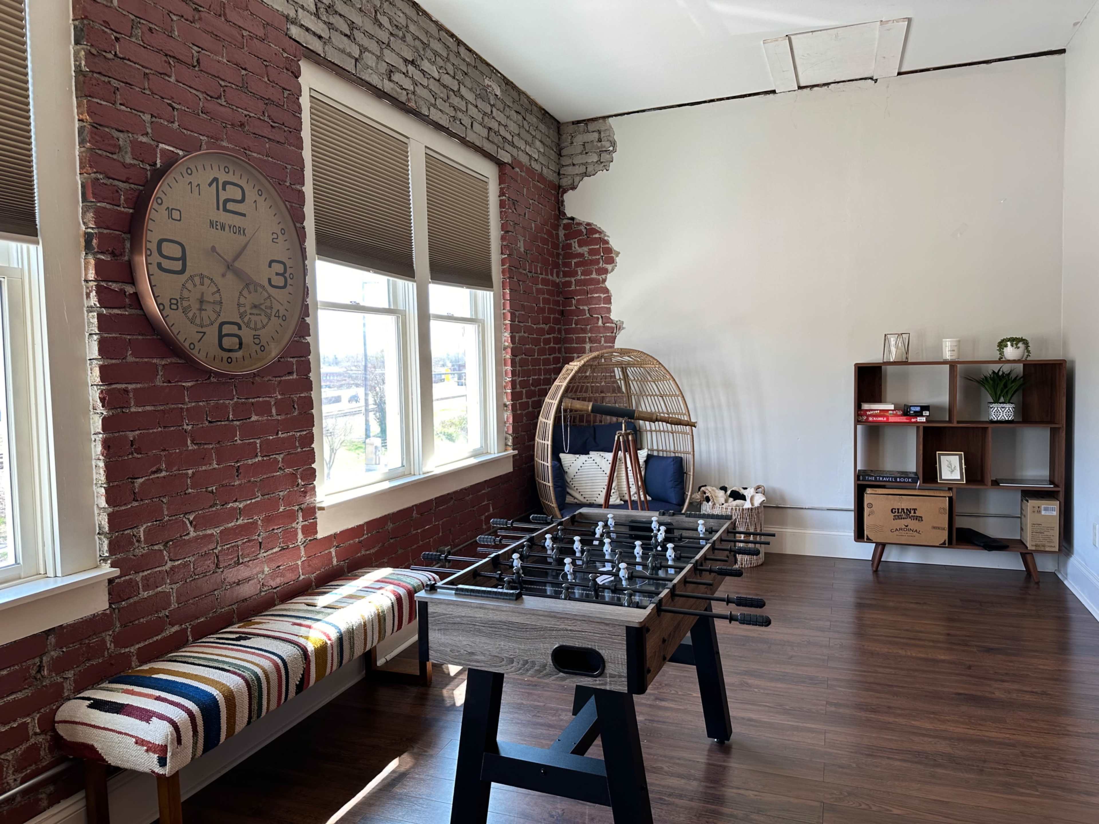 The scene features a room with exposed brick walls, a large clock, a foosball table, a striped bench, and a bookshelf.