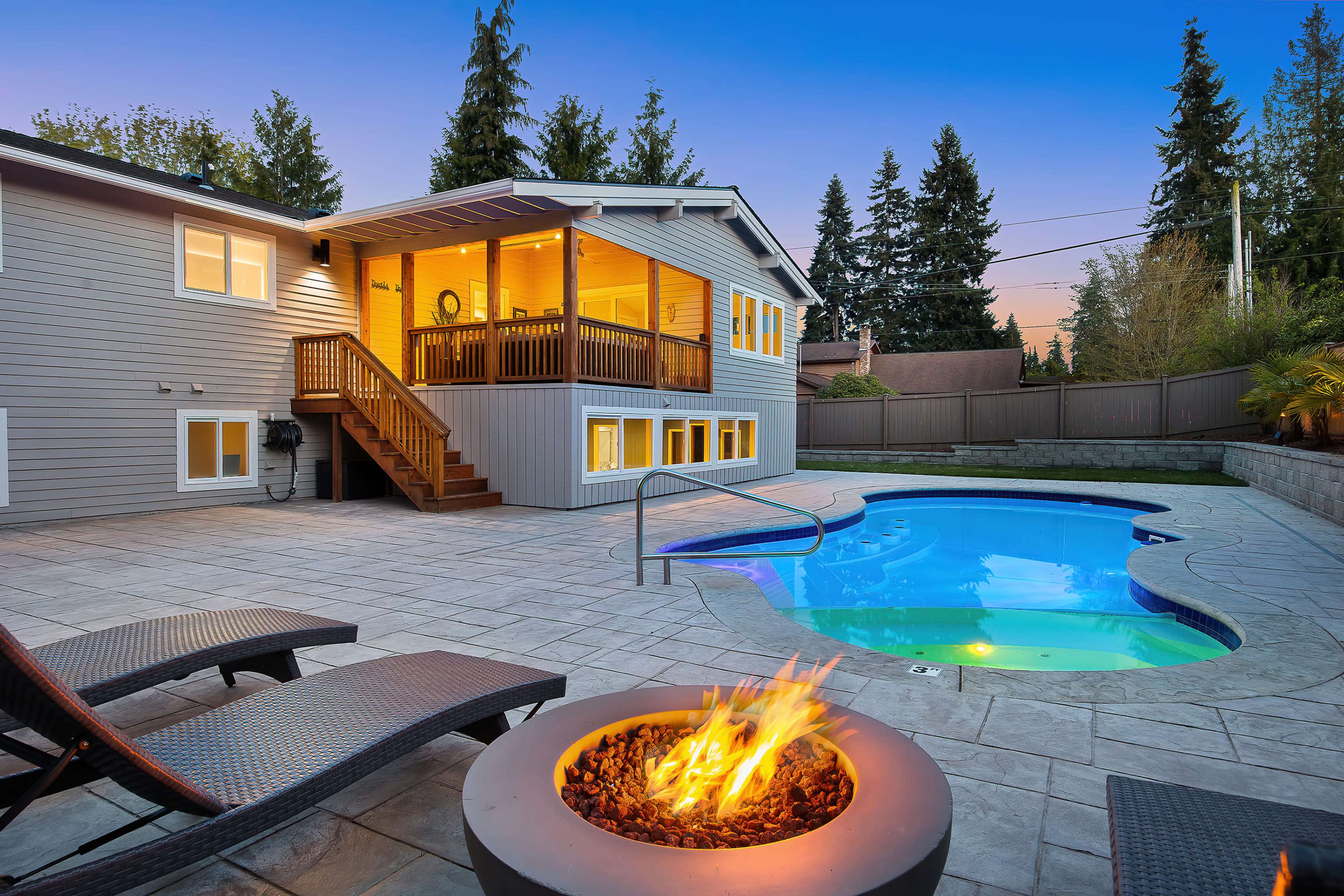 A backyard features a swimming pool, a fire pit, and a two-level house with a deck overlooking the area.