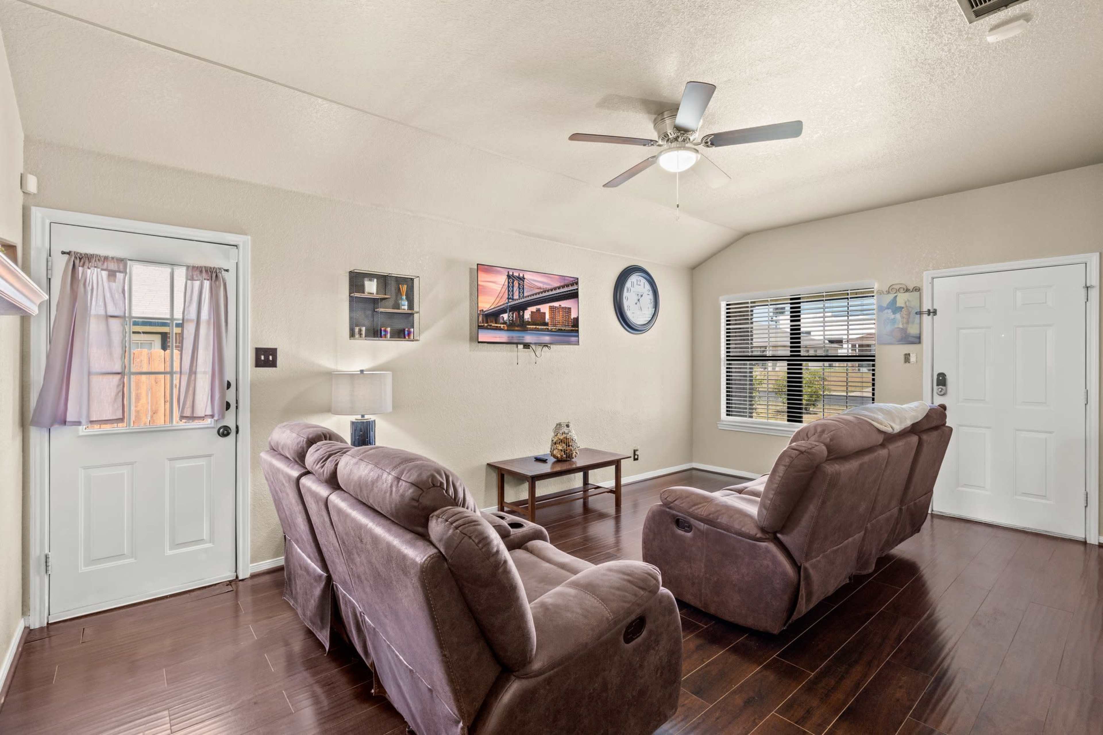 The living room features two reclining chairs facing a wall-mounted television, with a door leading outside and a clock on the wall.