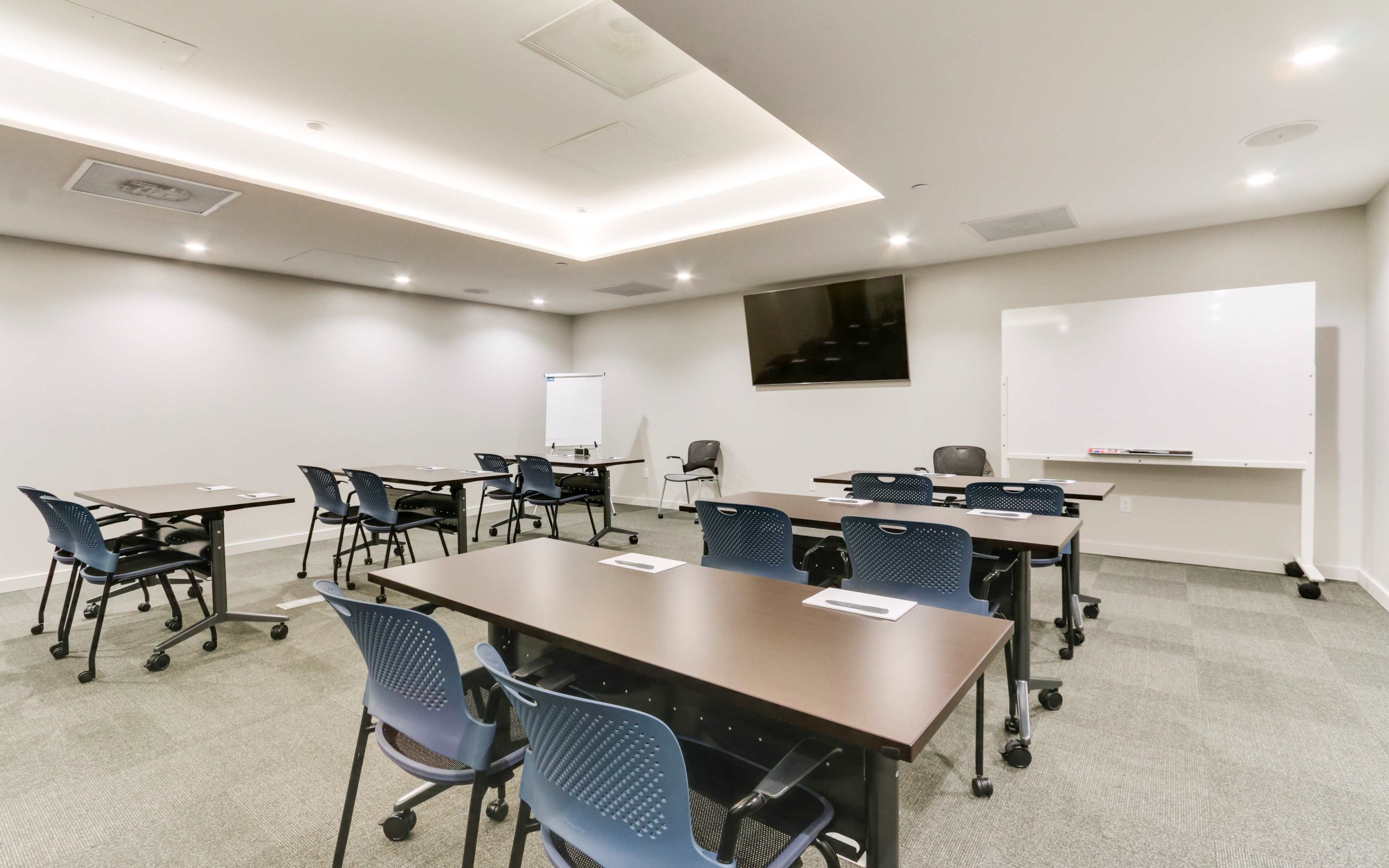 A modern conference room features several tables with chairs arranged in a U-shape, a whiteboard, and a large screen mounted on the wall.