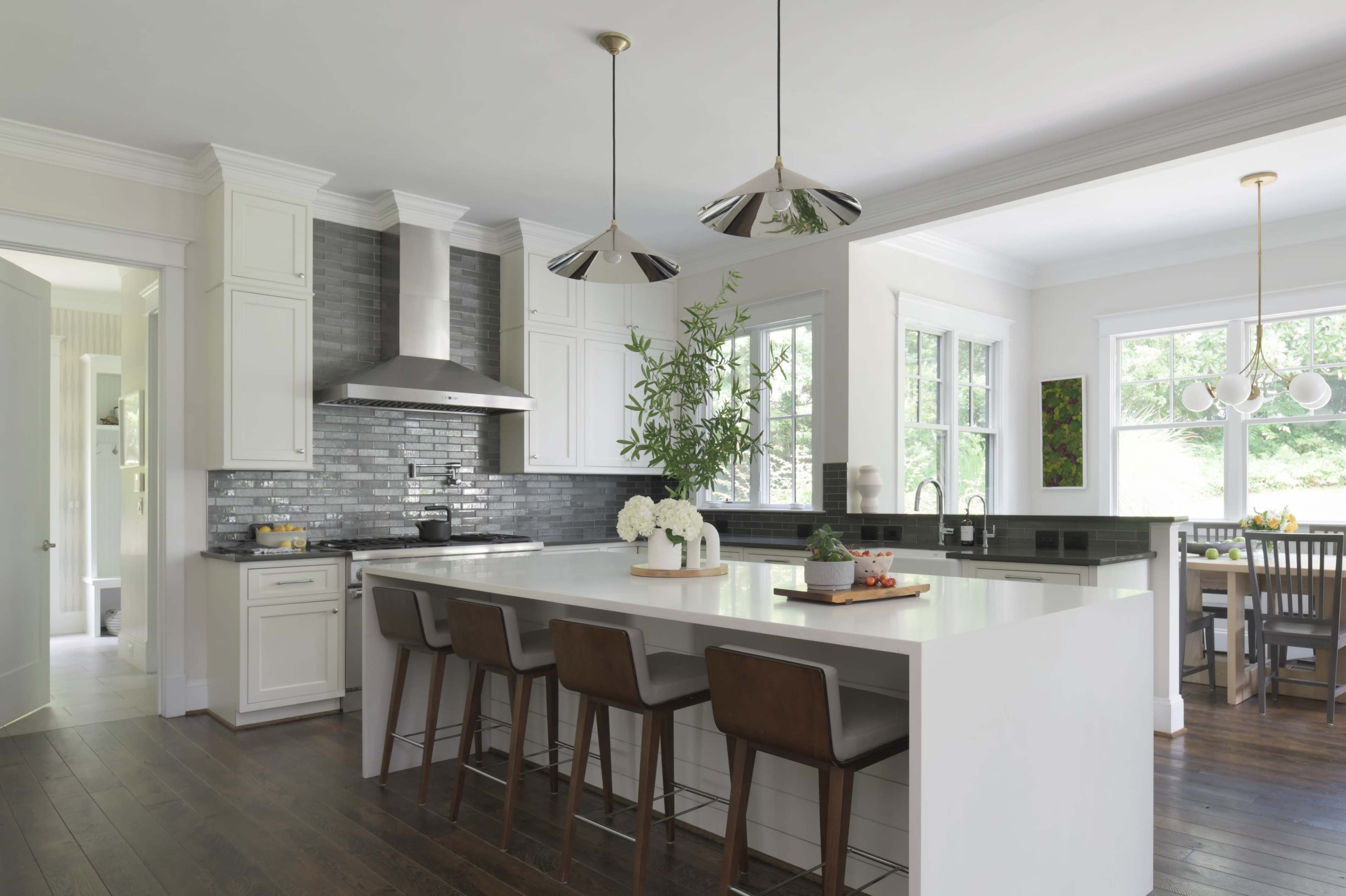 A modern kitchen features a large white island with four brown stools, stainless steel appliances, and natural light streaming through the windows.