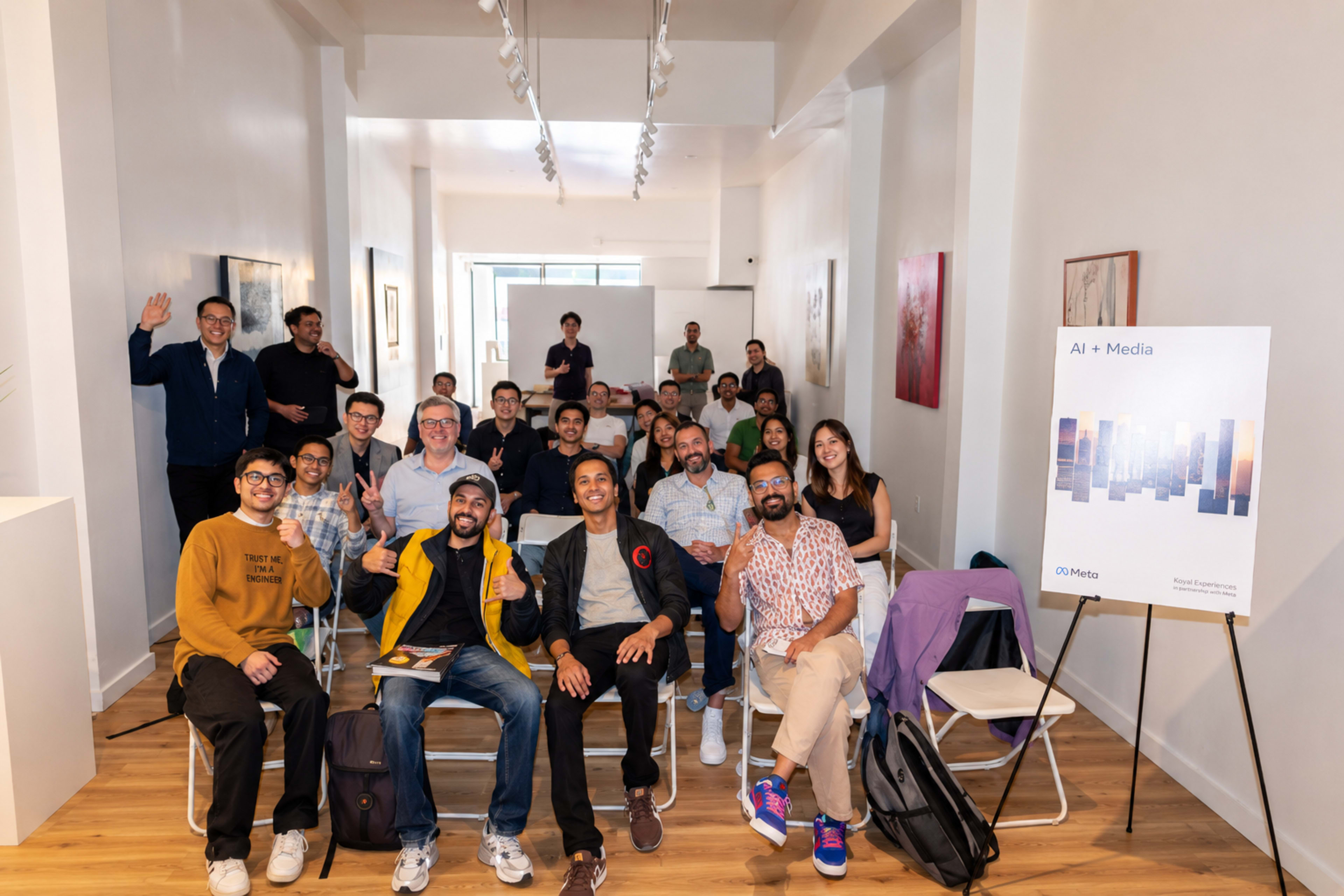 A diverse group of individuals is gathered in a well-lit room, posing for a photo with some waving and a sign displaying "AI + Media" in the background.