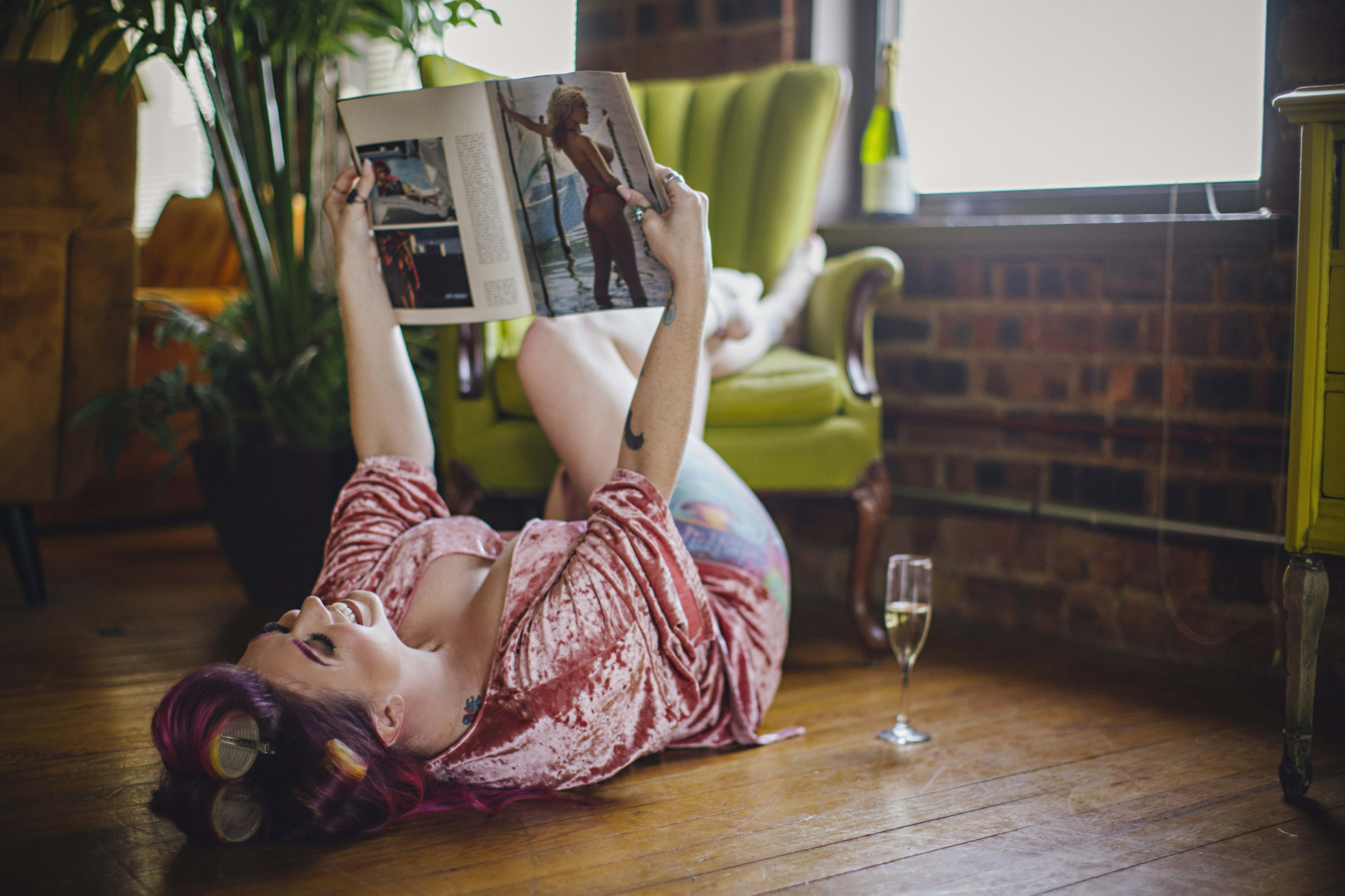 A woman lounging on a rustic brown floor amidst green plants, engrossed in a magazine.