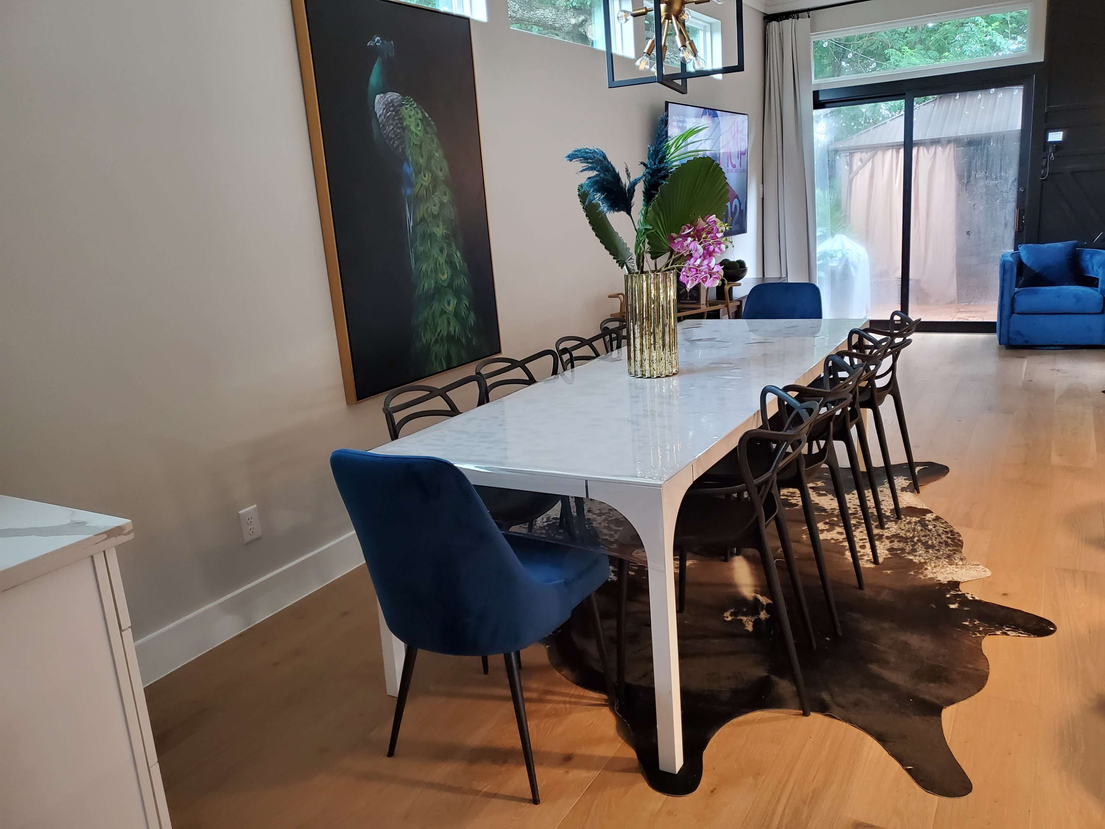 A modern dining room features a long white marble table surrounded by black chairs and a decorative vase, with a peacock painting on the wall and a blue sofa in the background.