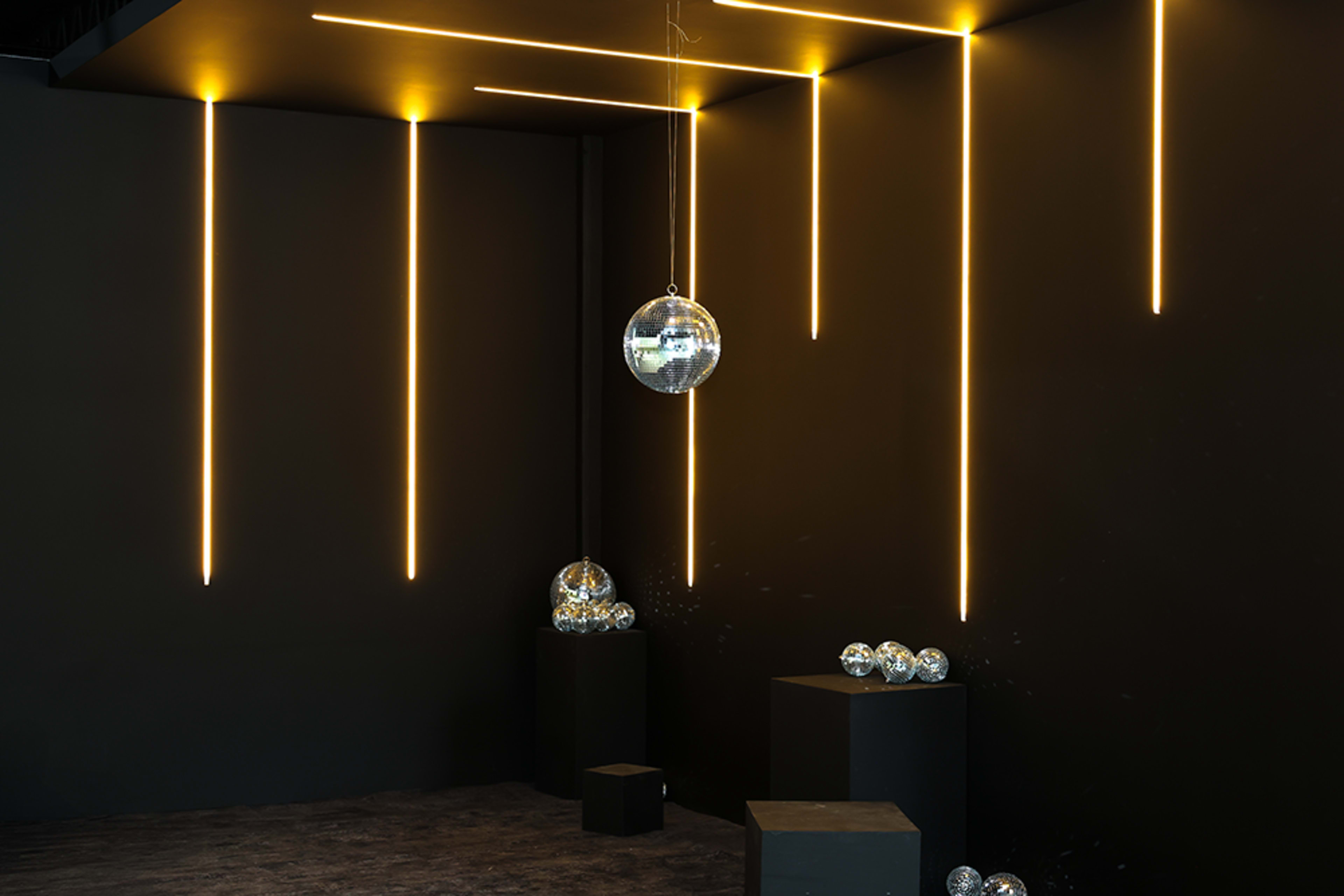 The image features a gallery space with a black background, illuminated by yellow LED lights, and adorned with multiple reflective disco balls placed on black pedestals.