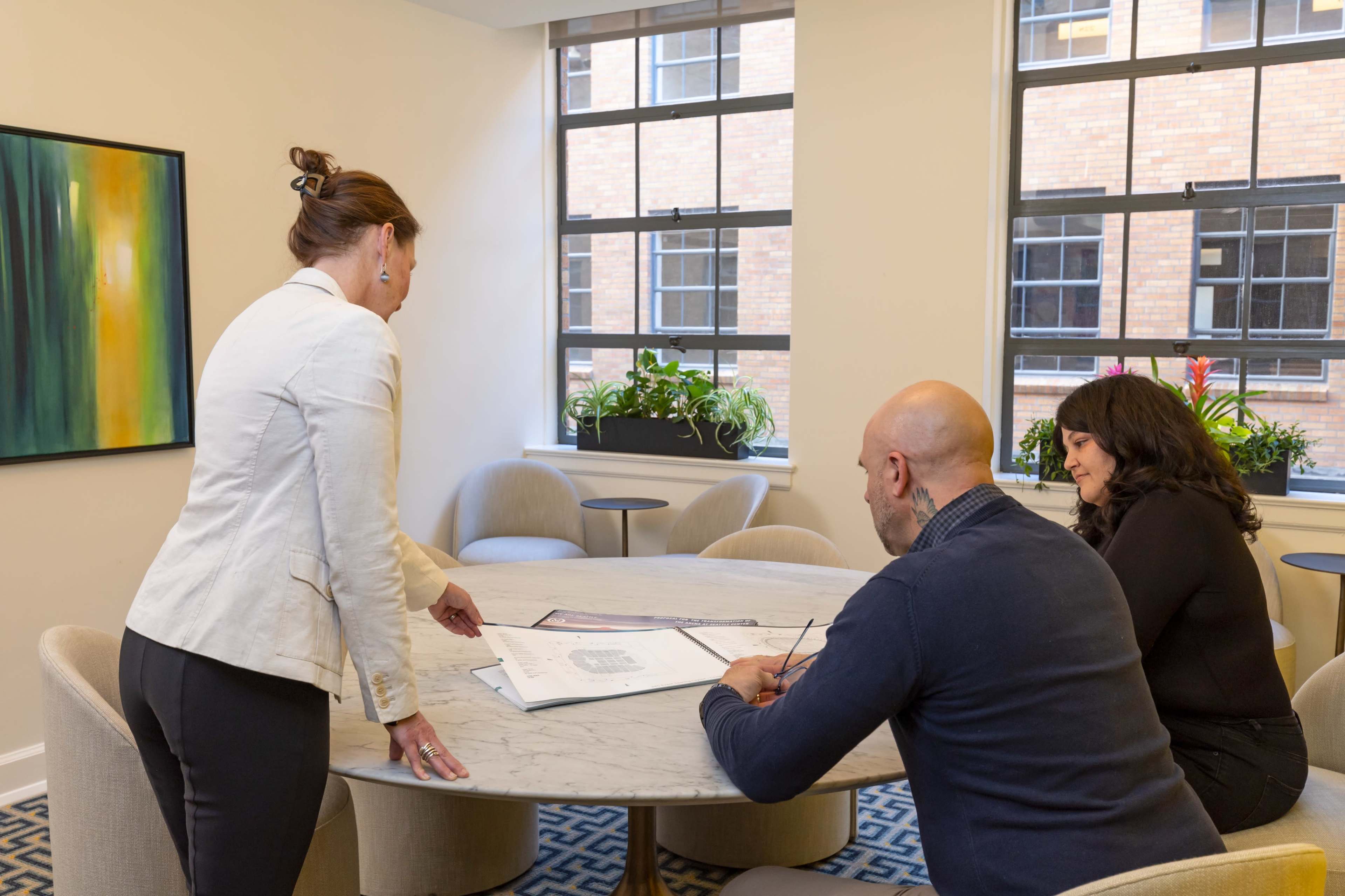 Three individuals are gathered around a marble conference table, discussing plans displayed on a document in the center.