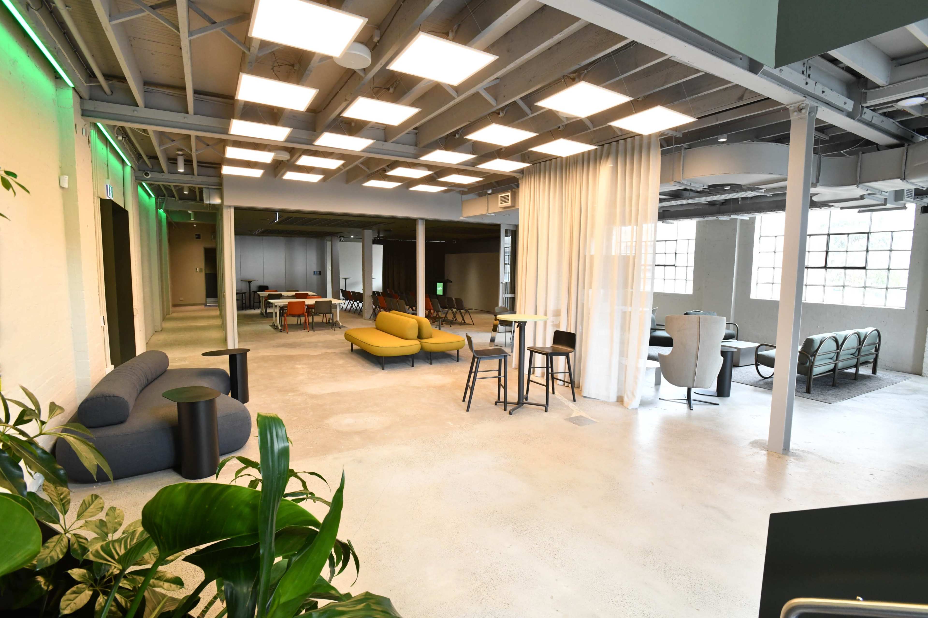 The image shows a spacious, modern interior with various seating arrangements and large windows, illuminated by overhead lights.