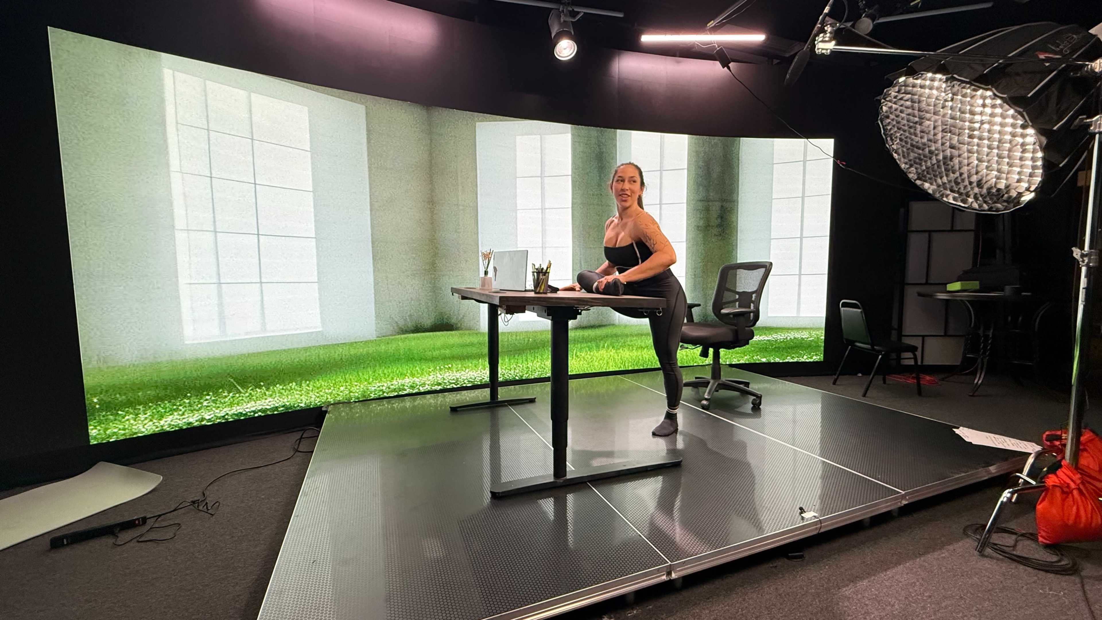A woman is seated on a desk in a studio with a large curved screen displaying a green grassy landscape and large windows.
