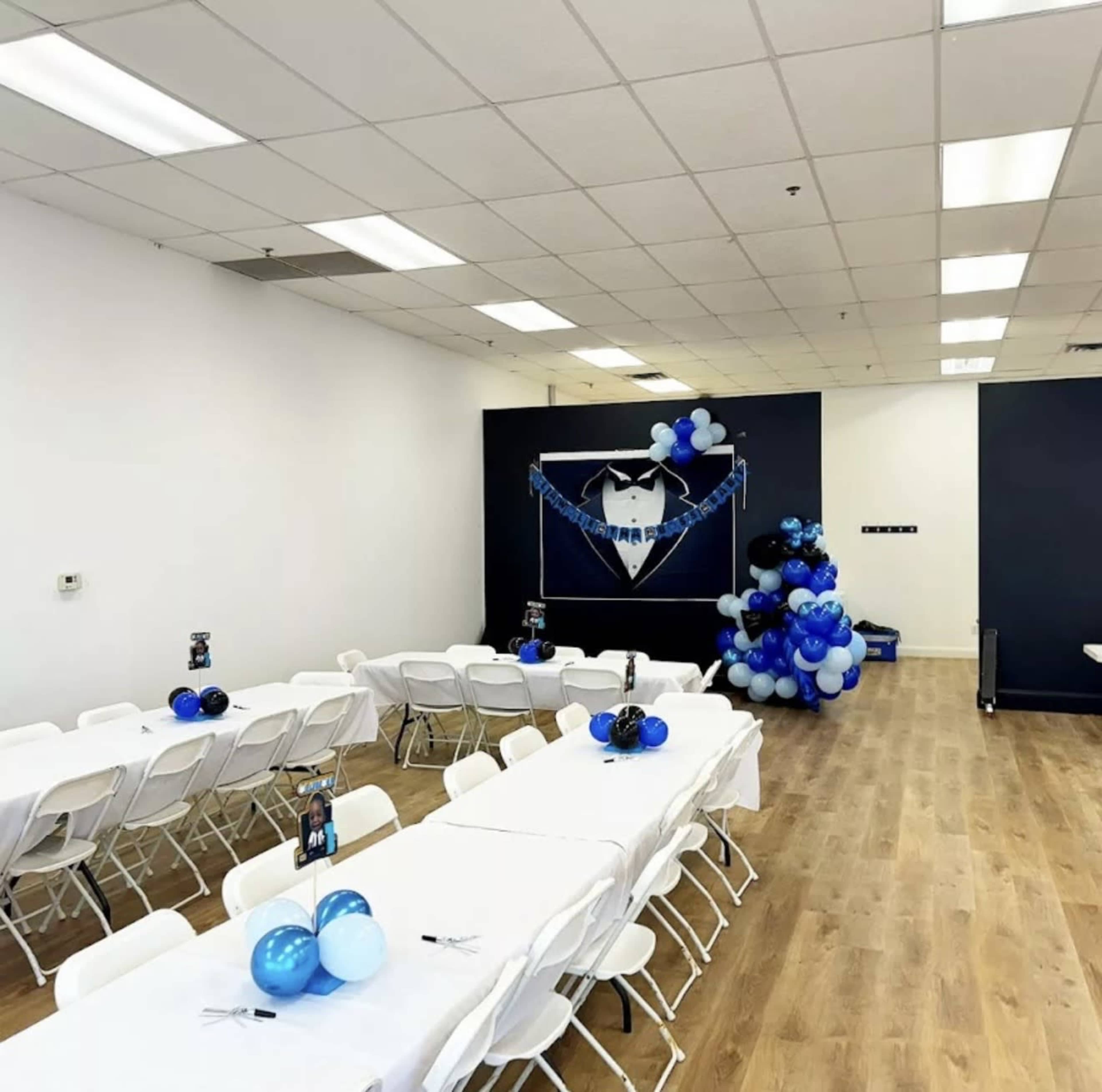 The scene shows a party venue with rows of white tables set with blue balloons and a decorated wall featuring a black and blue theme.