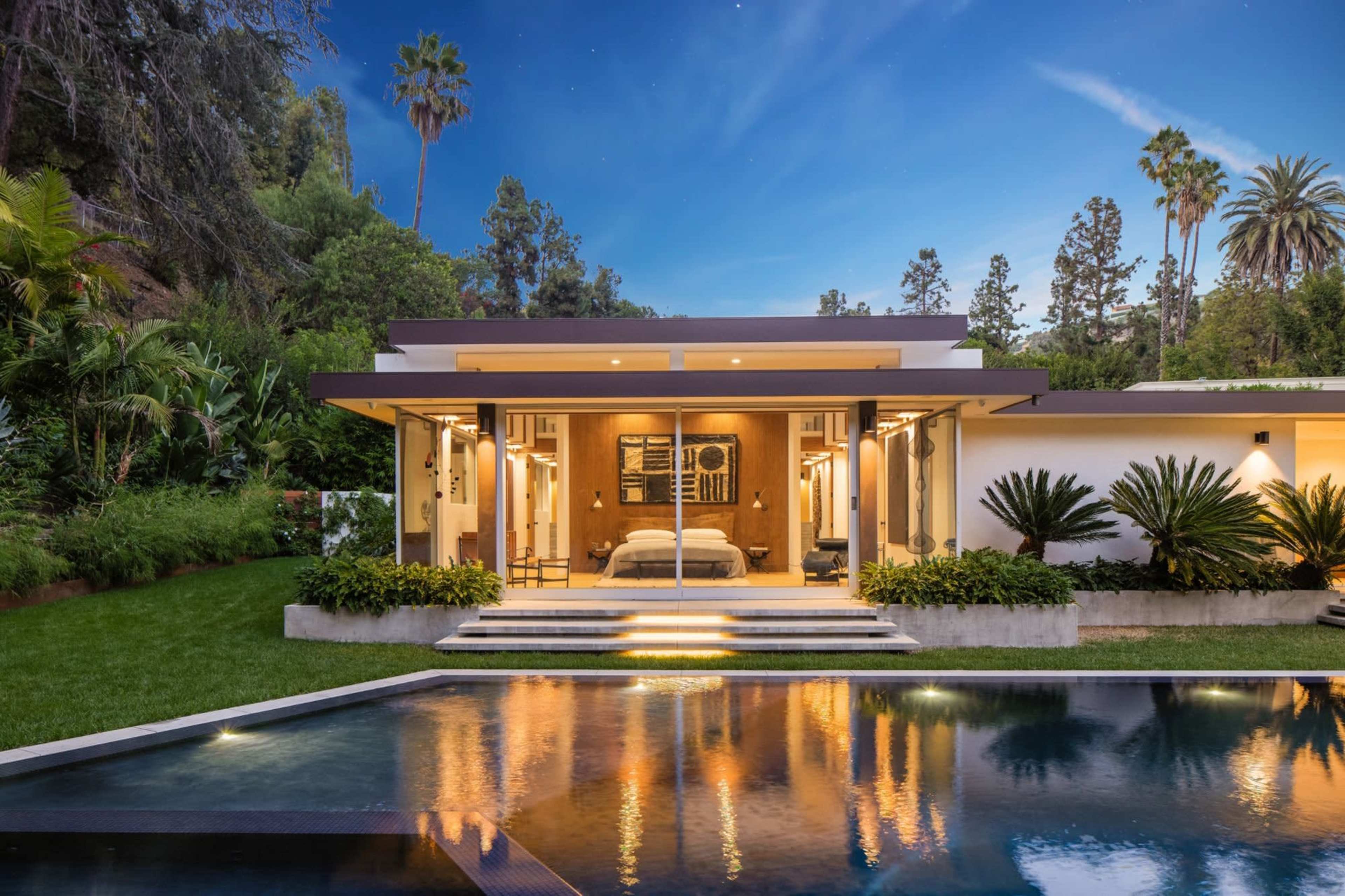 A modern house with a large glass sliding door opens to a patio overlooking a swimming pool and lush greenery under a twilight sky.