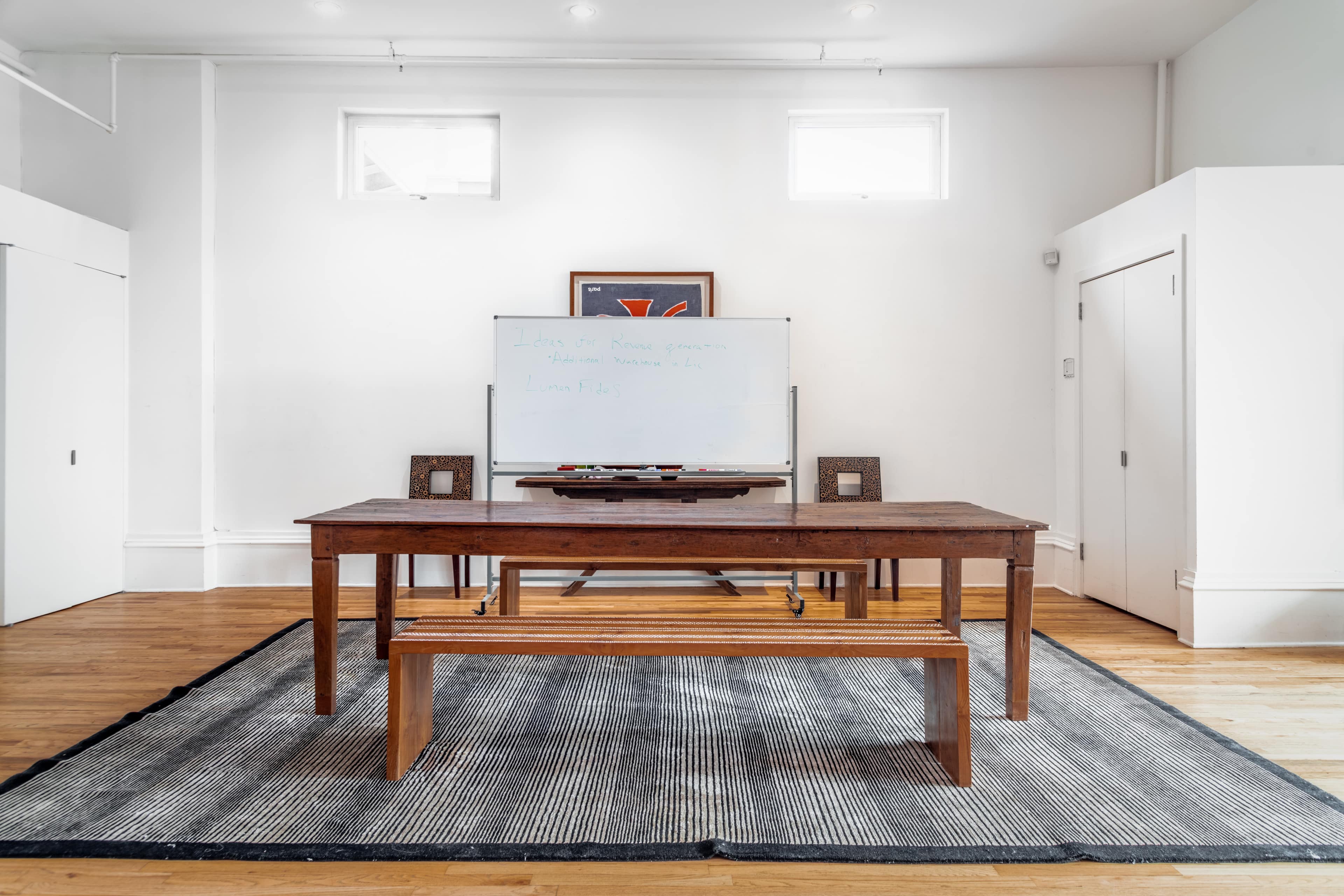 A wooden table and bench are set on a striped rug in a bright, minimalist room with a whiteboard and artwork on the walls.