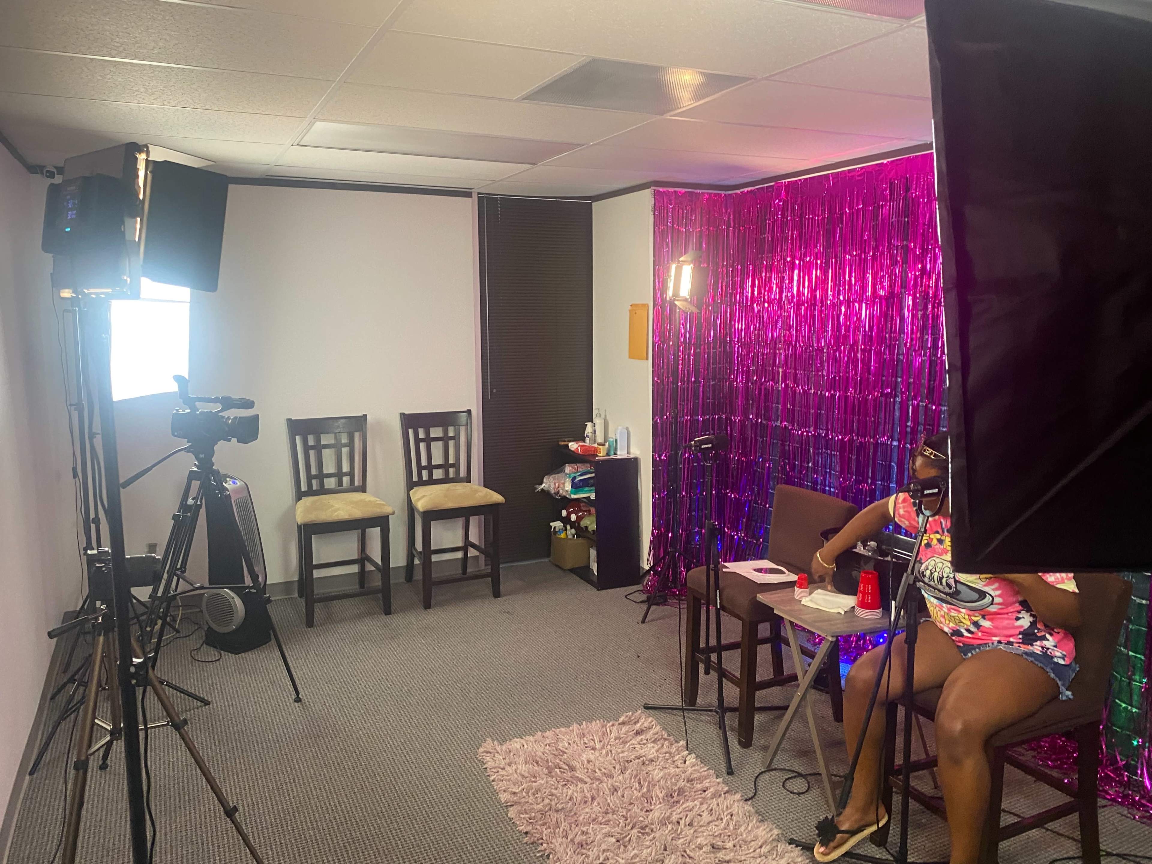 A small studio space is set up with cameras, lights, and two chairs, featuring a pink metallic background and a table with various items.