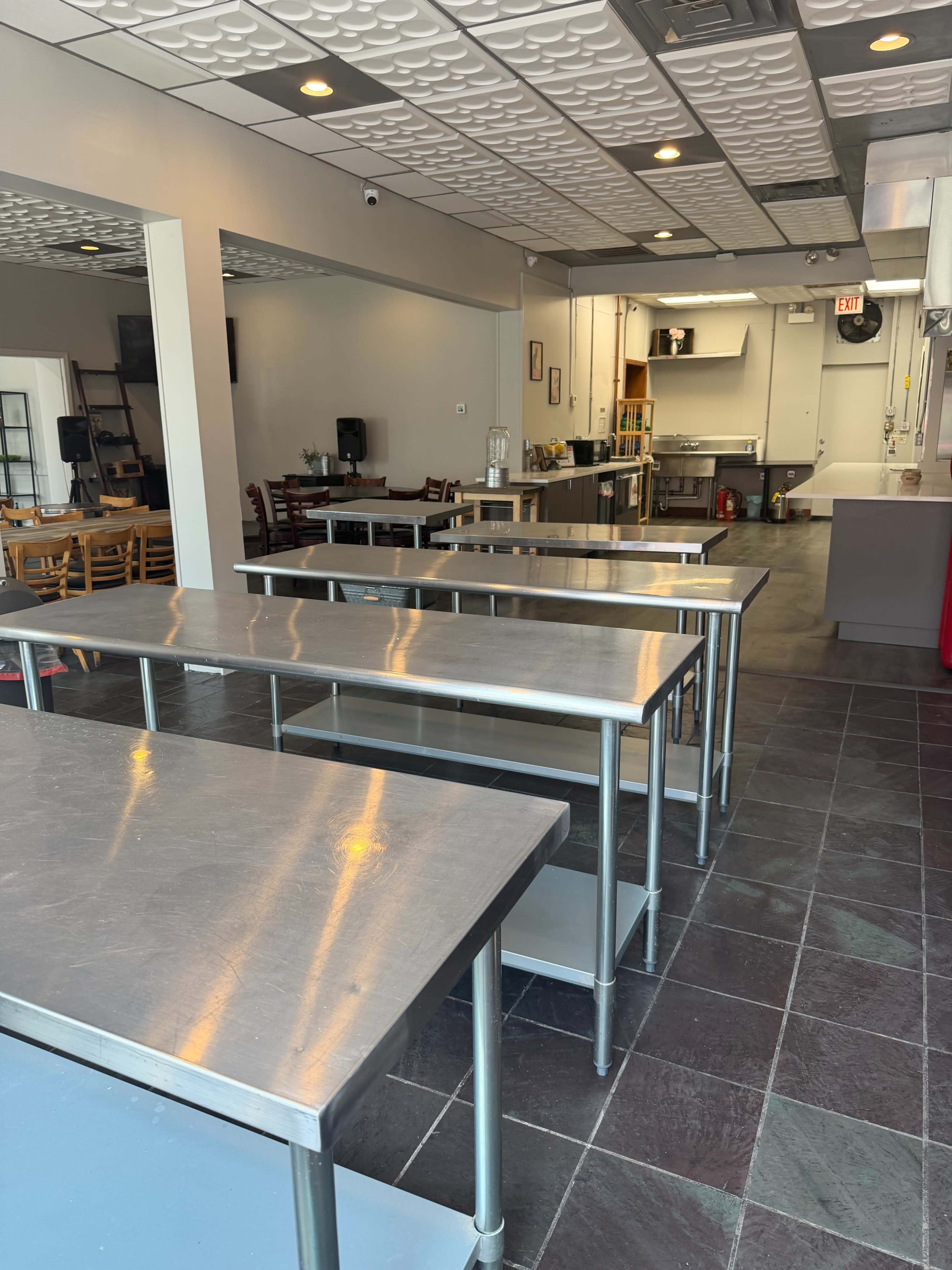 The image shows a clean, spacious restaurant interior with stainless steel tables, a visible kitchen area, and a few empty chairs.