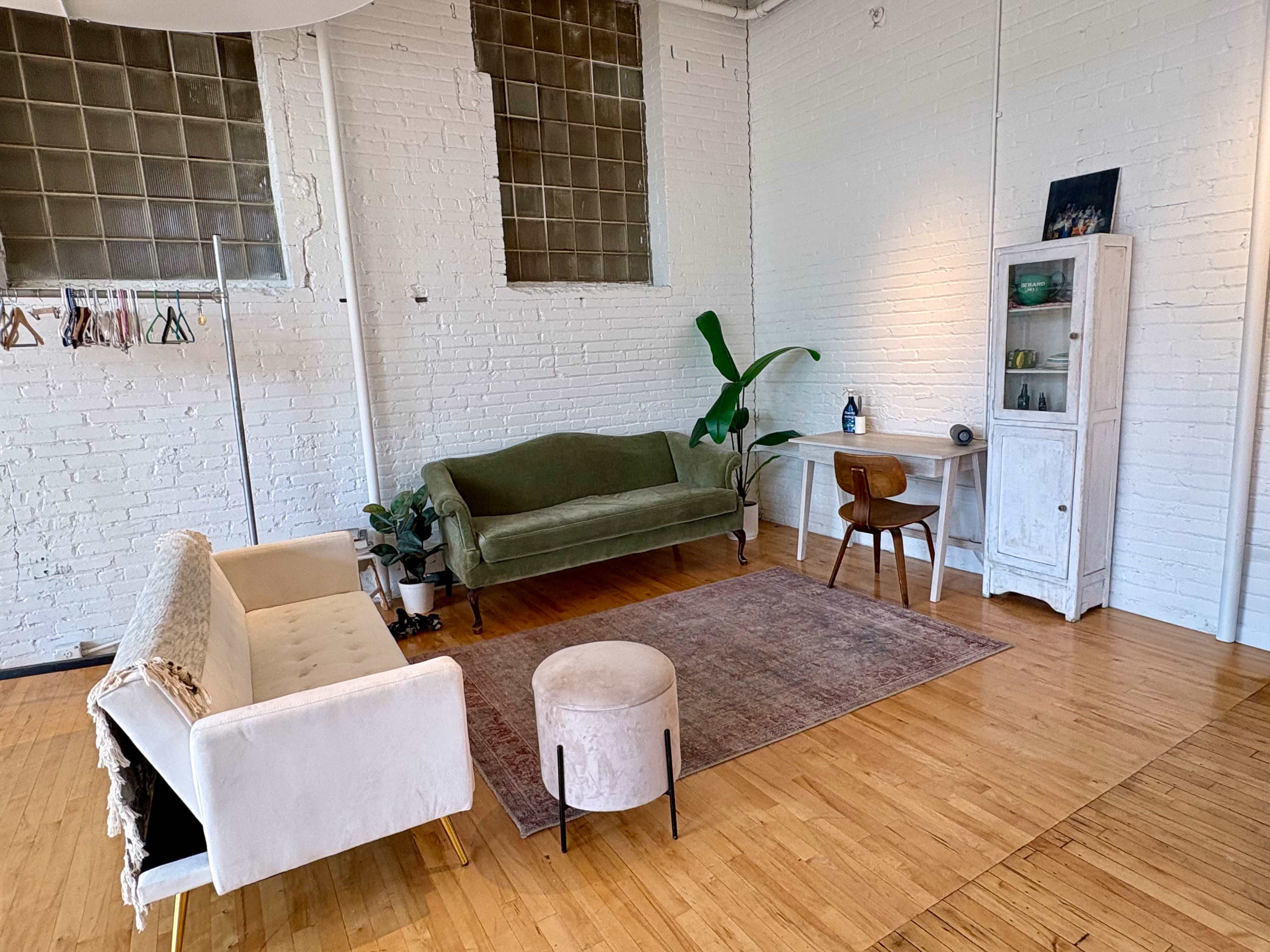 The image shows a well-lit, minimalist interior space featuring a green sofa, a small white table, a wooden chair, a pouf, and a plant against a brick wall.
