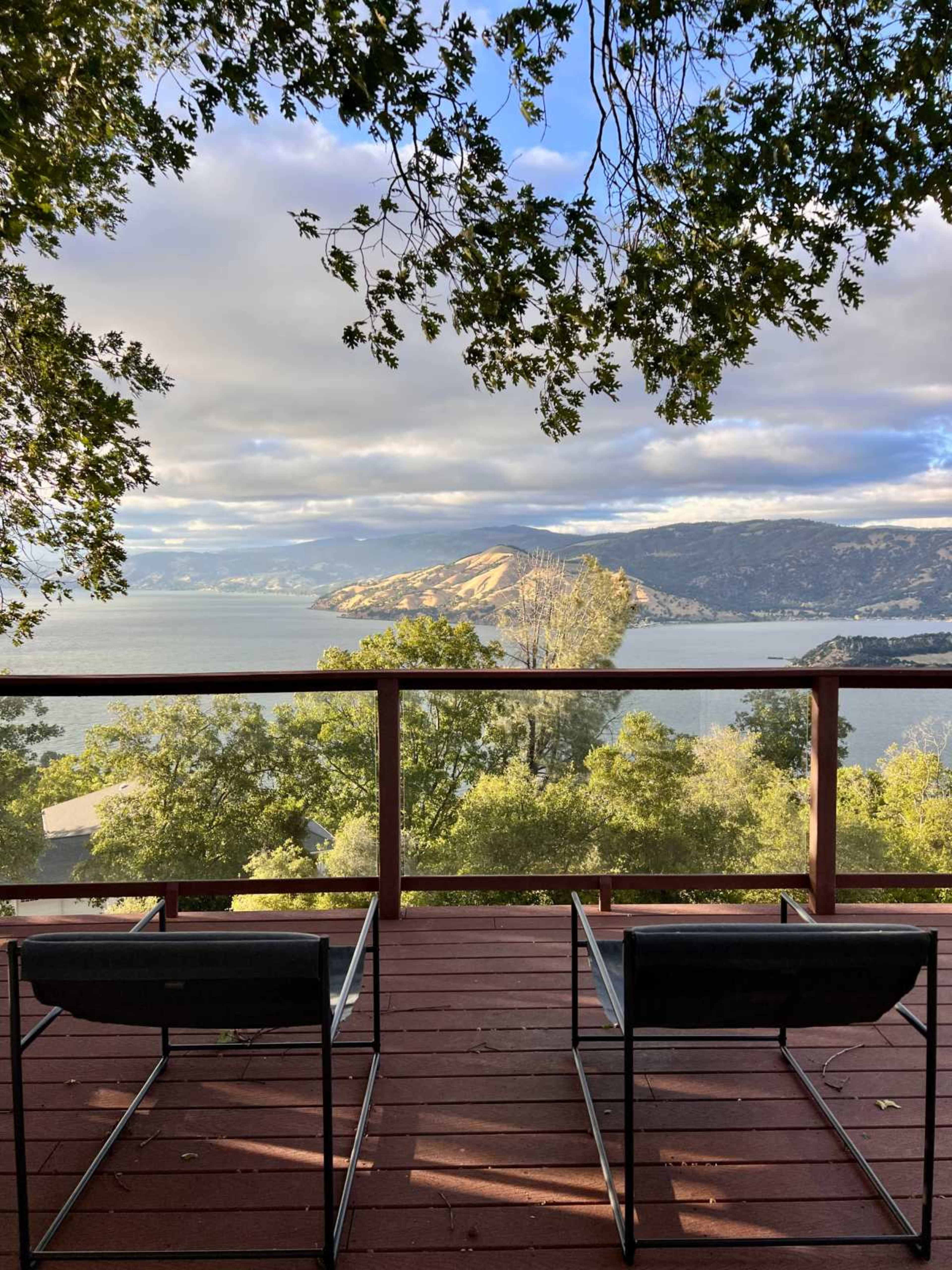 A wooden deck with two chairs overlooks a lake and a mountainous shoreline under a mostly cloudy sky.